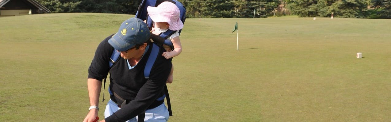 A father golfs while wearing a baby in a backpack-style carrier on his back. He wears a baseball cap and the baby wears a pink gingham bucket hat.