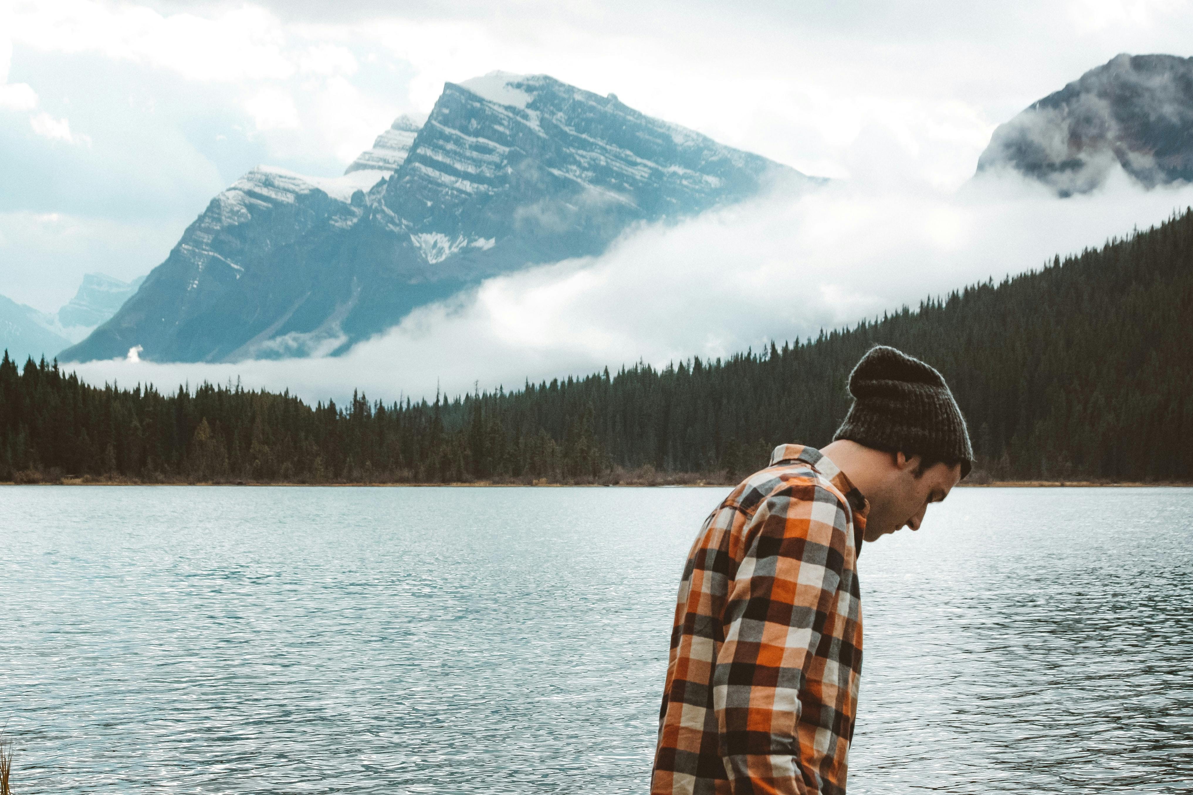 A man in a plaid shirt walks along a lake with a mountain and trees in the distance