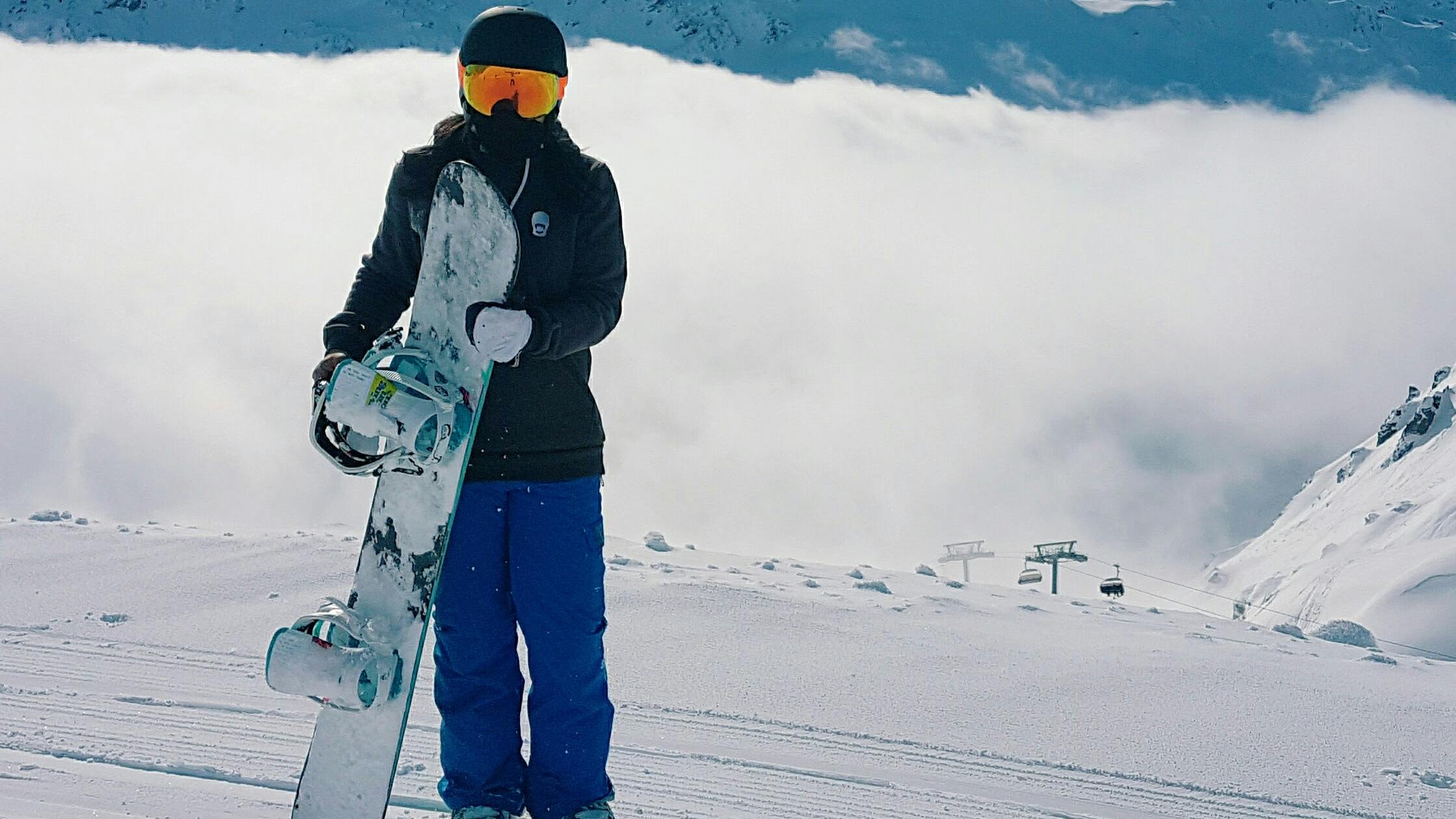 A snowboarder in a black jacket and blue pants looks at the camera