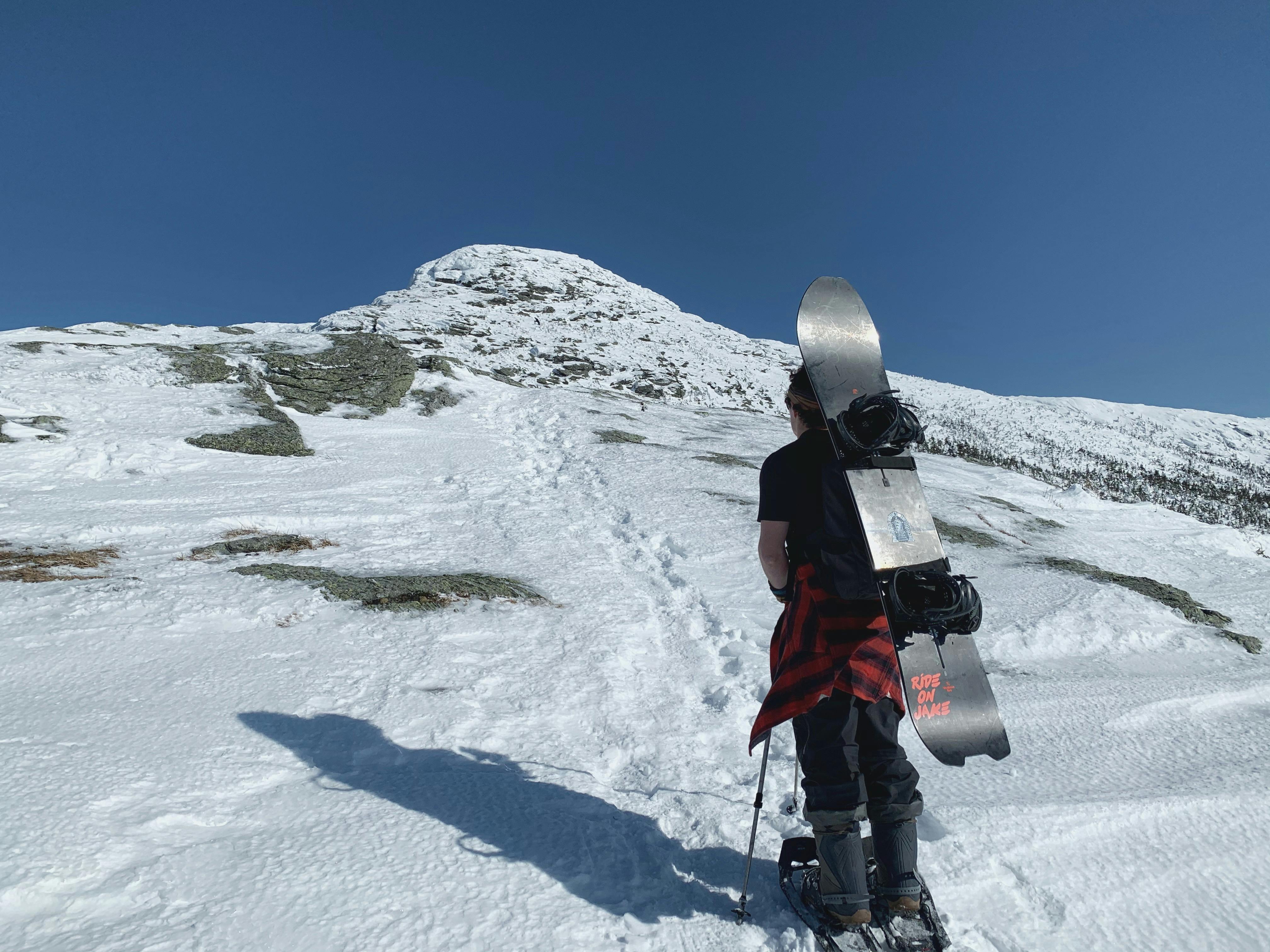 A person wears their snowboard on their back as they scale a hill in snowshoes