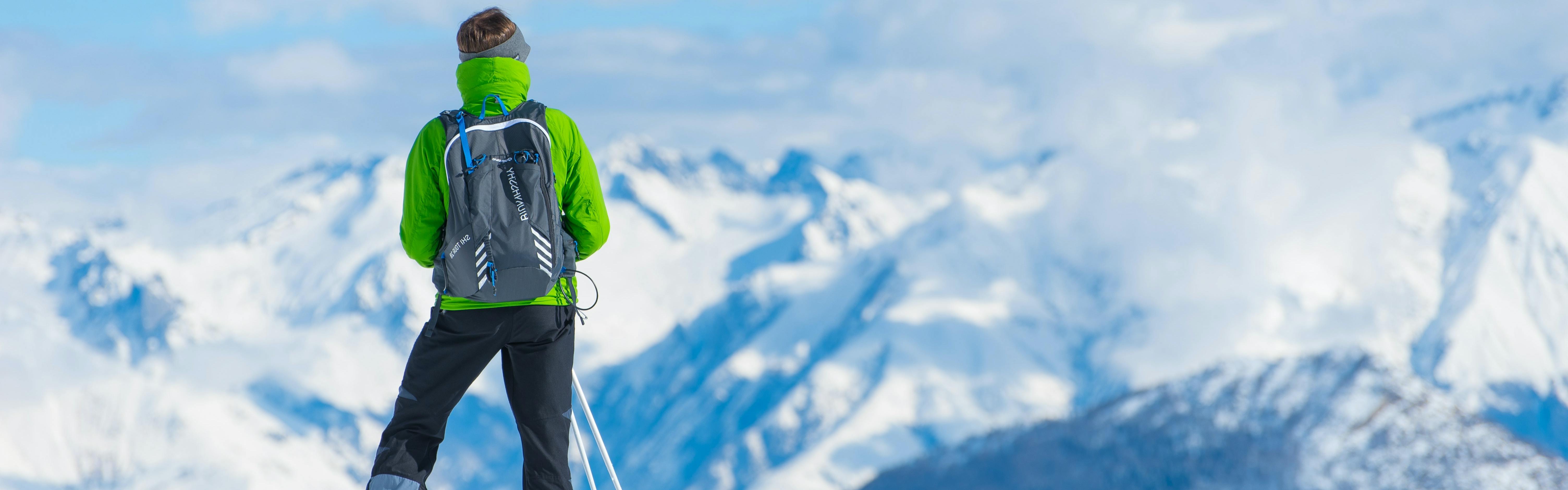A skier wearing a green jacket and a grey backpack stands at the top of a ski pass. It is sunny and you can see a lot of snow covered peaks in the background.