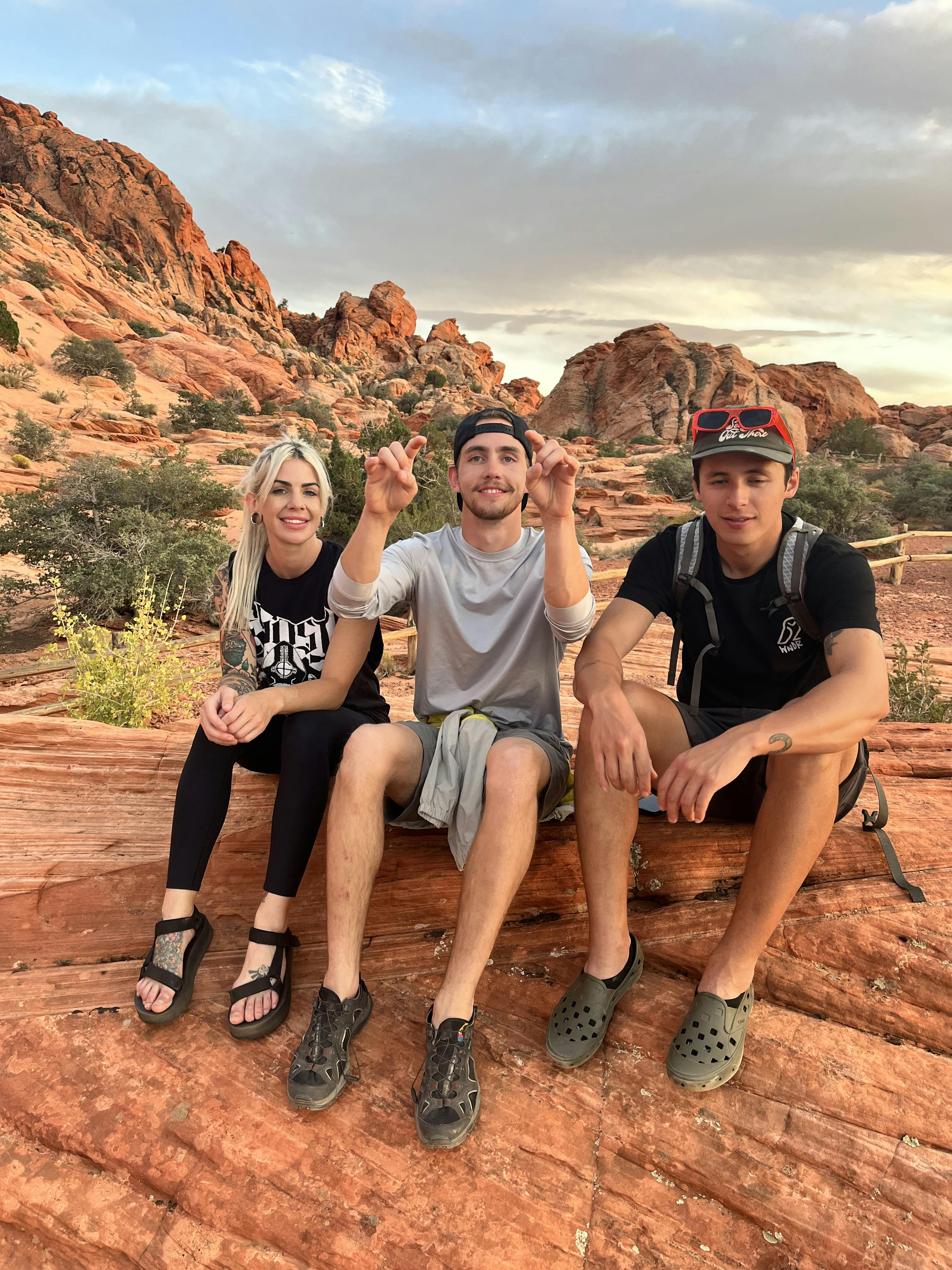 Three hikers sitting together. Two of them are wearing sandals. There are red rocks in the background.