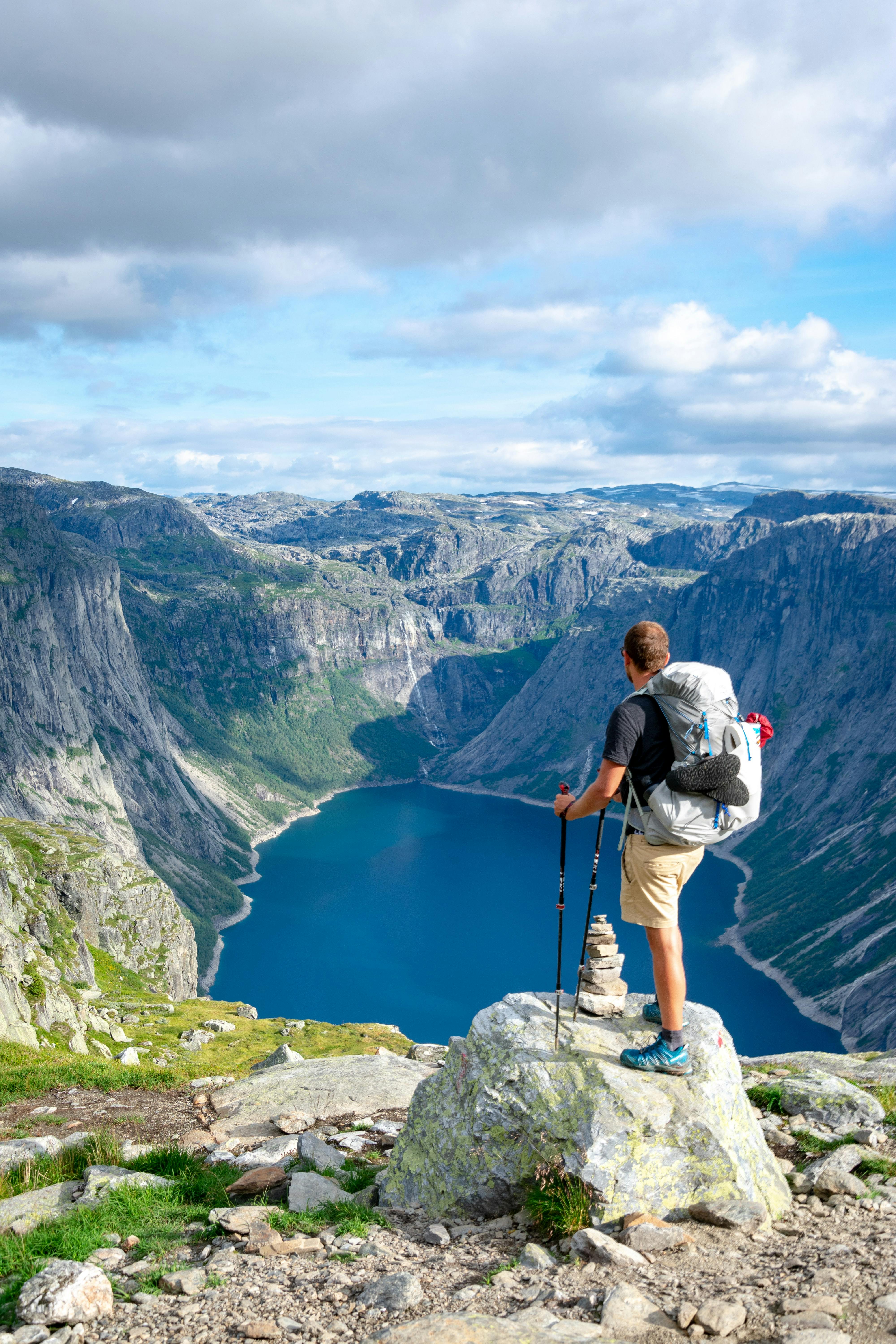 A man stands on a rock sitting on a peak that overlooks a wide valley filled with a lake. He's in an alpine environment with rocky terrain and low-lying plant life. He wears trail runners.