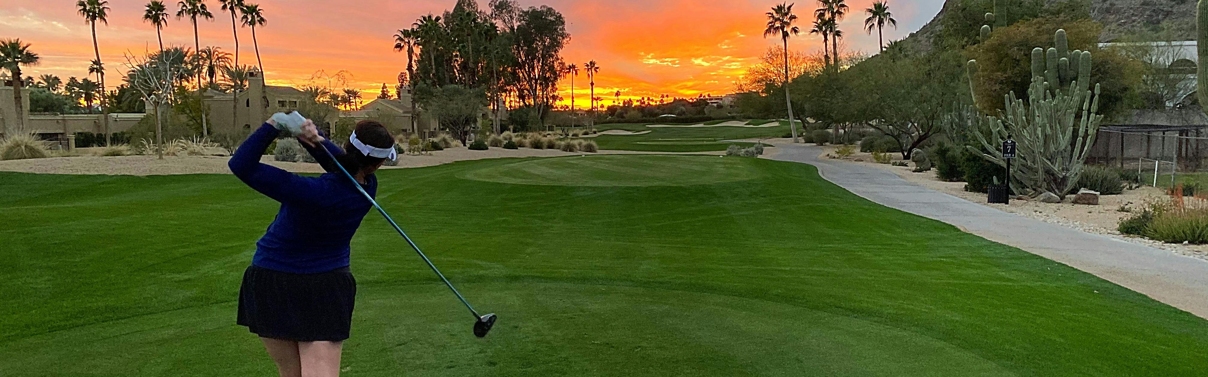 A woman swings a golf club while looking out towards an orange sunset