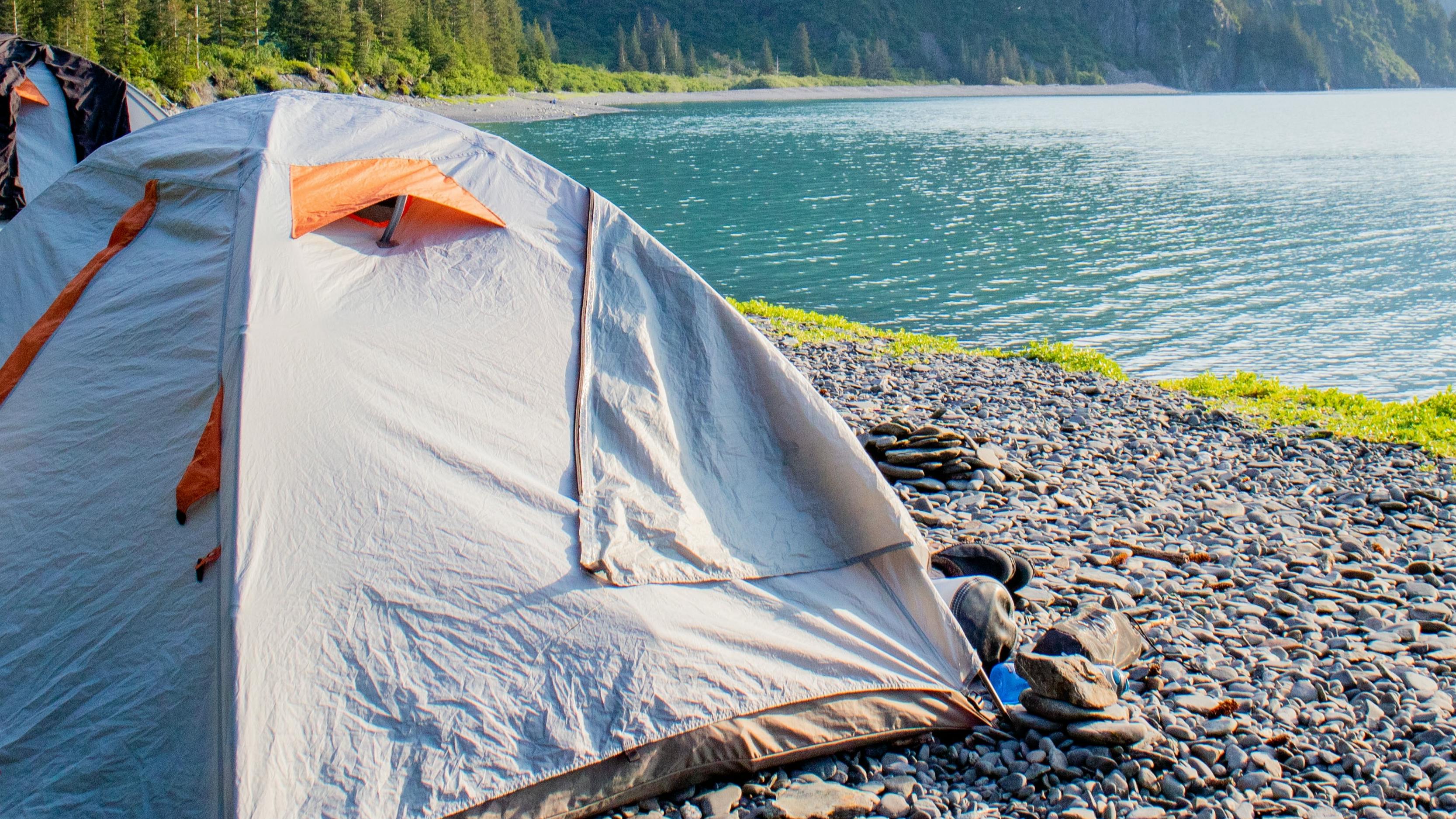 Two grey and orange tents are pitched by the edge of a lake. There are some pants drying on top of one of the tents.