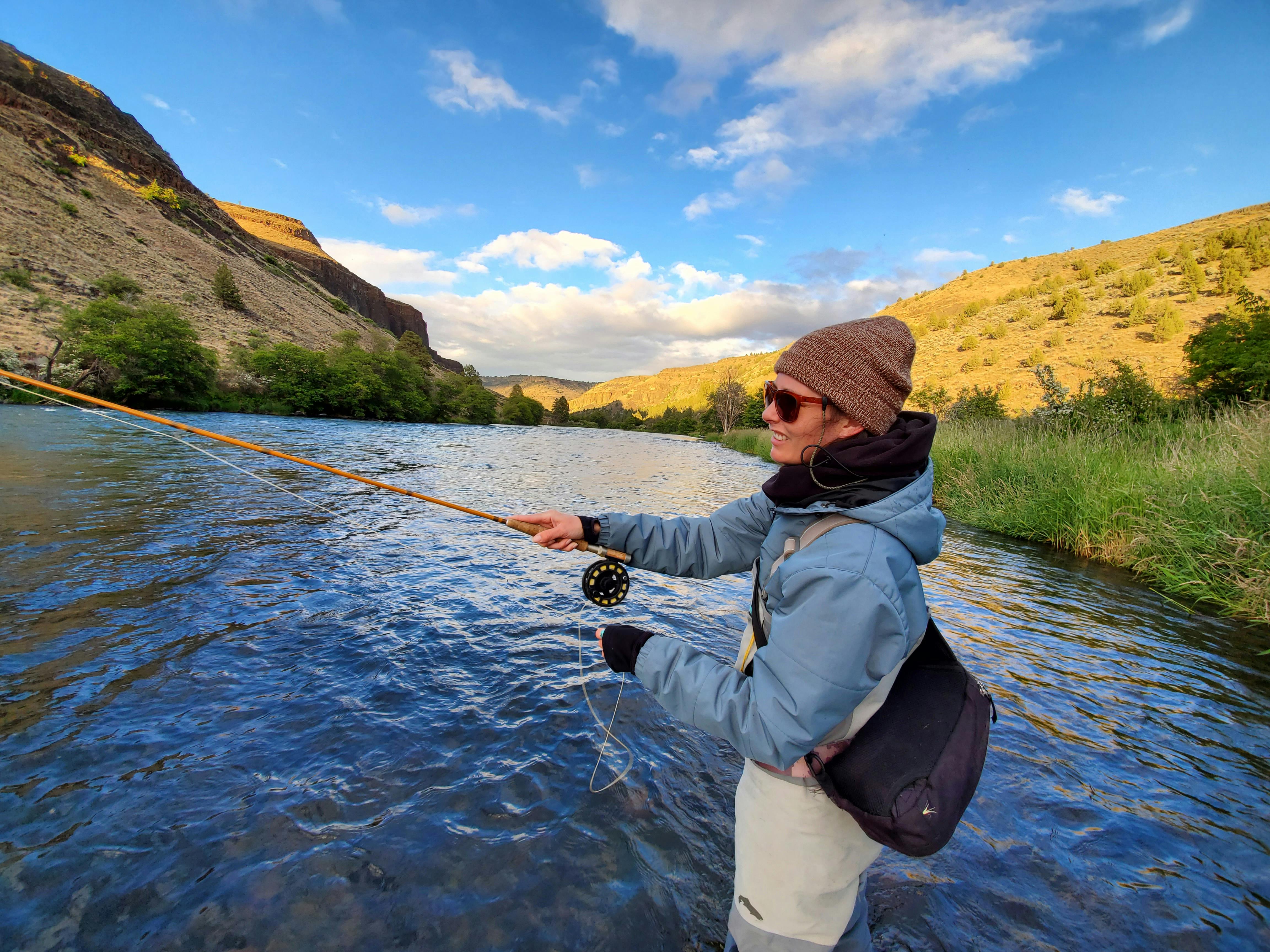A woman in a hat, a blue jacket, and waders casts a fly fishing rod