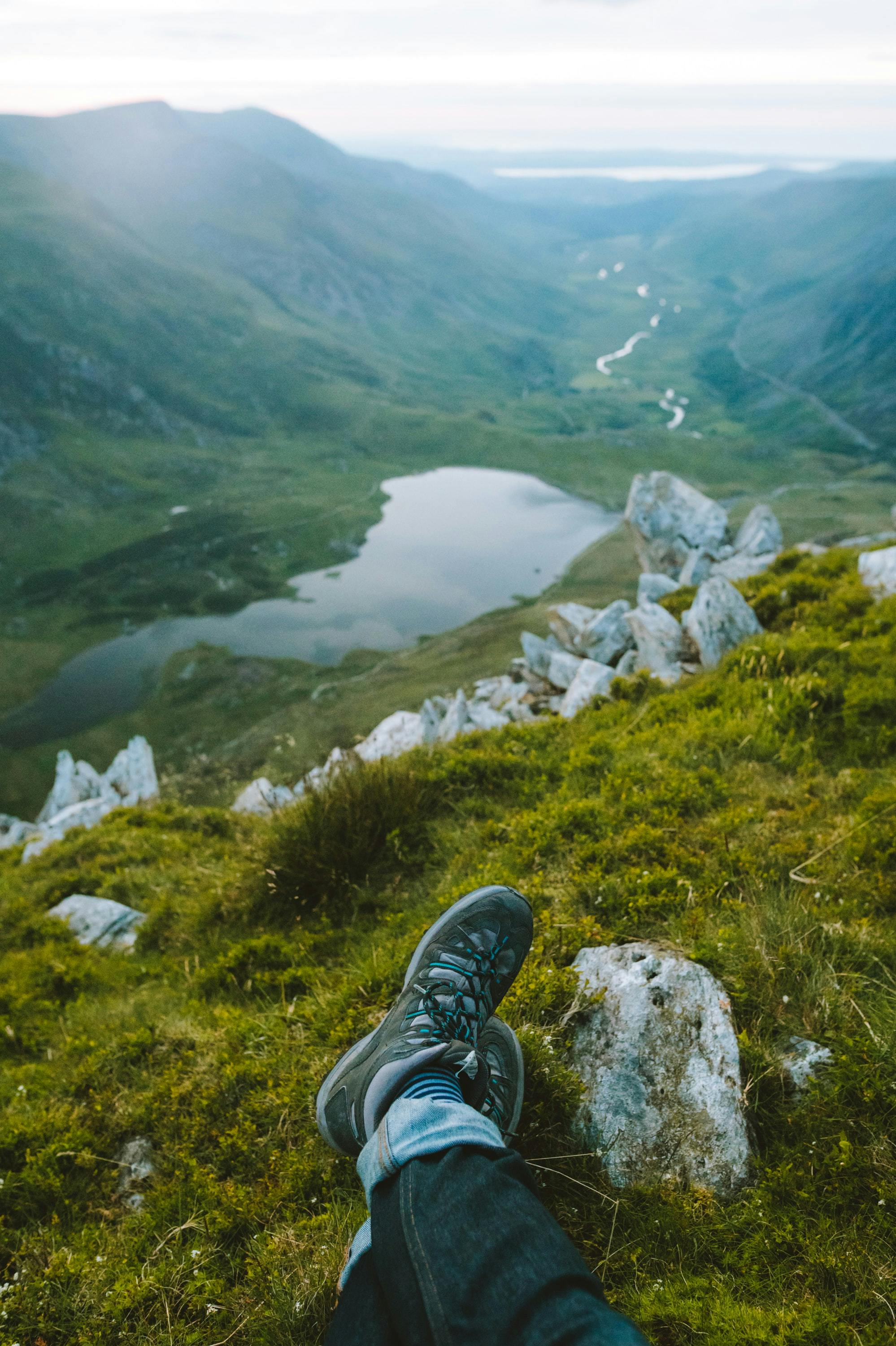 A person stretches out their legs as they sit overlooking a lake and mountains