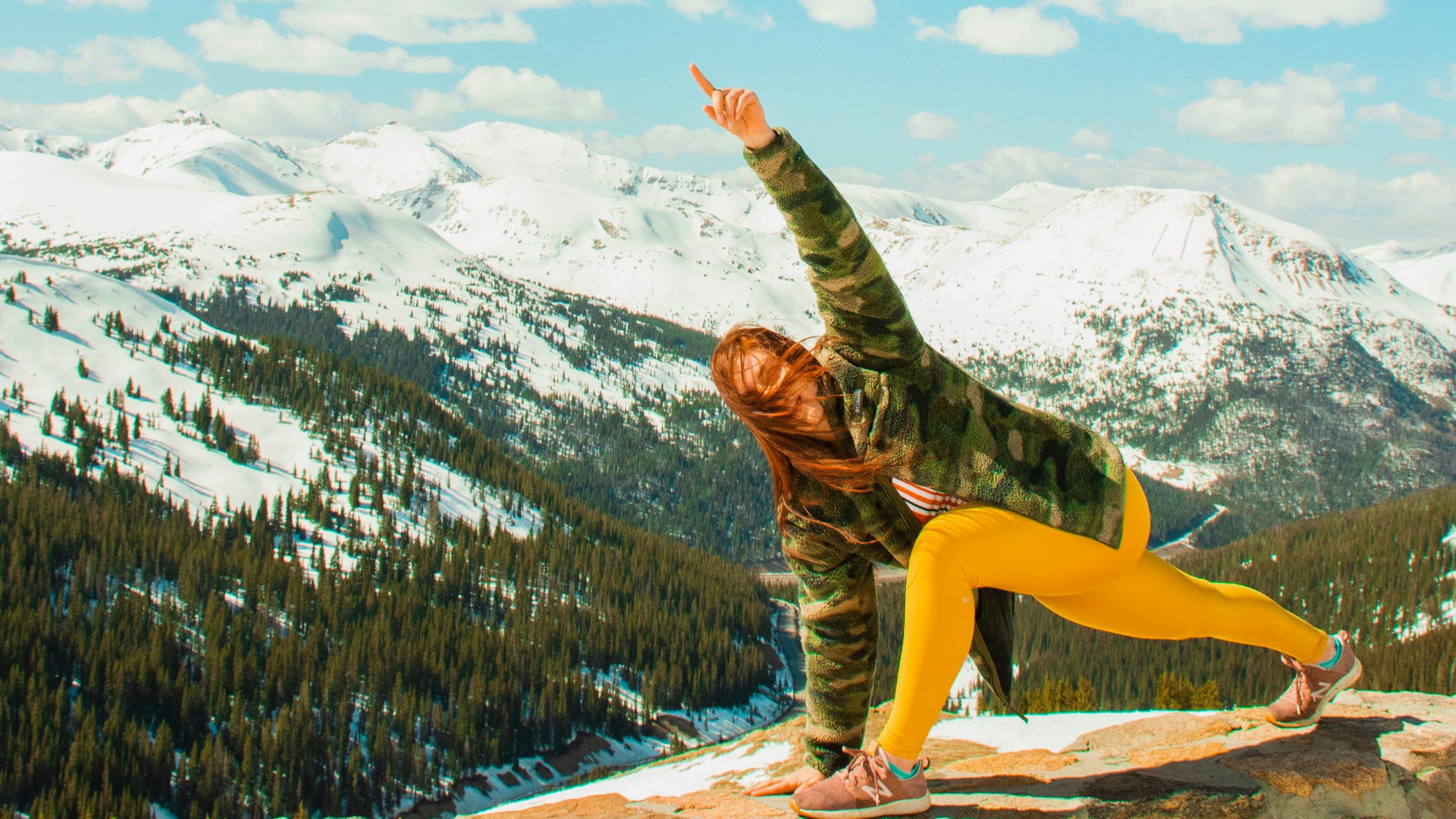 Woman does yoga on the top of a peak. There are snowy mountains in the background.