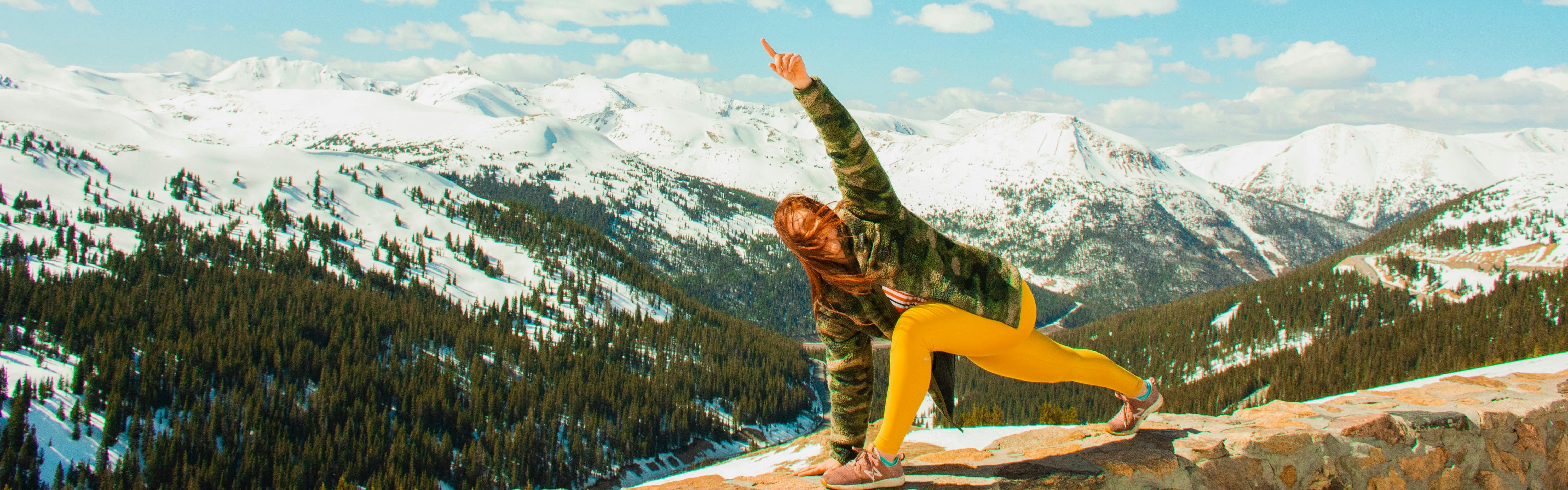 Woman does yoga on the top of a peak. There are snowy mountains in the background.