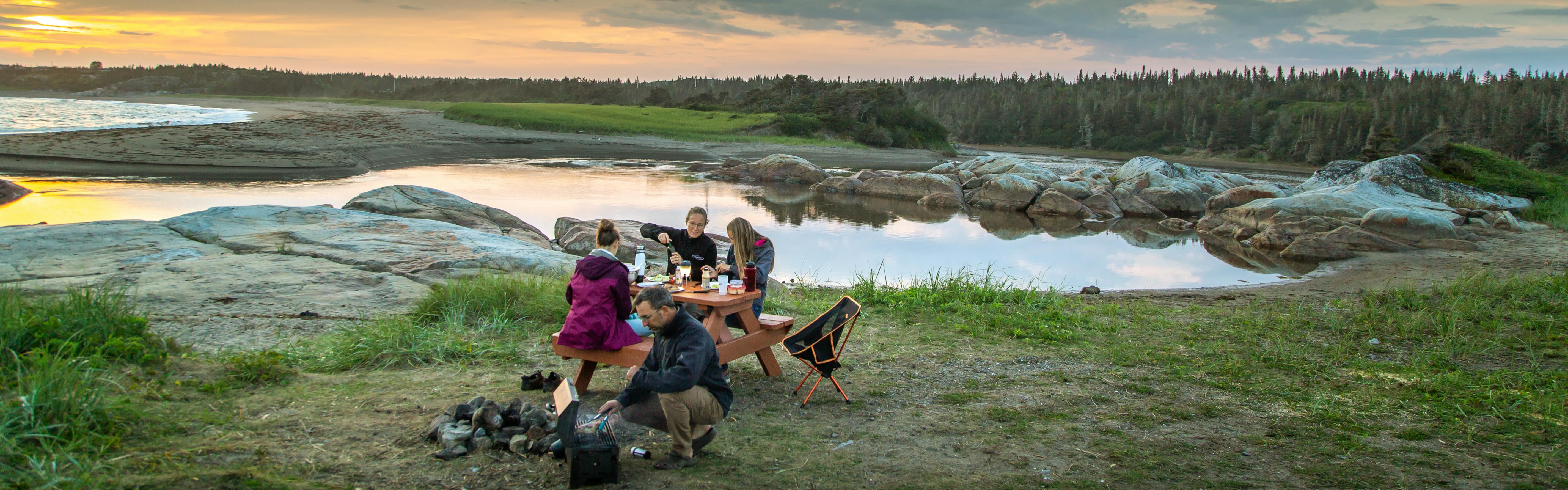 A family sits at a lake-side picnic table at sunset and shares a meal.