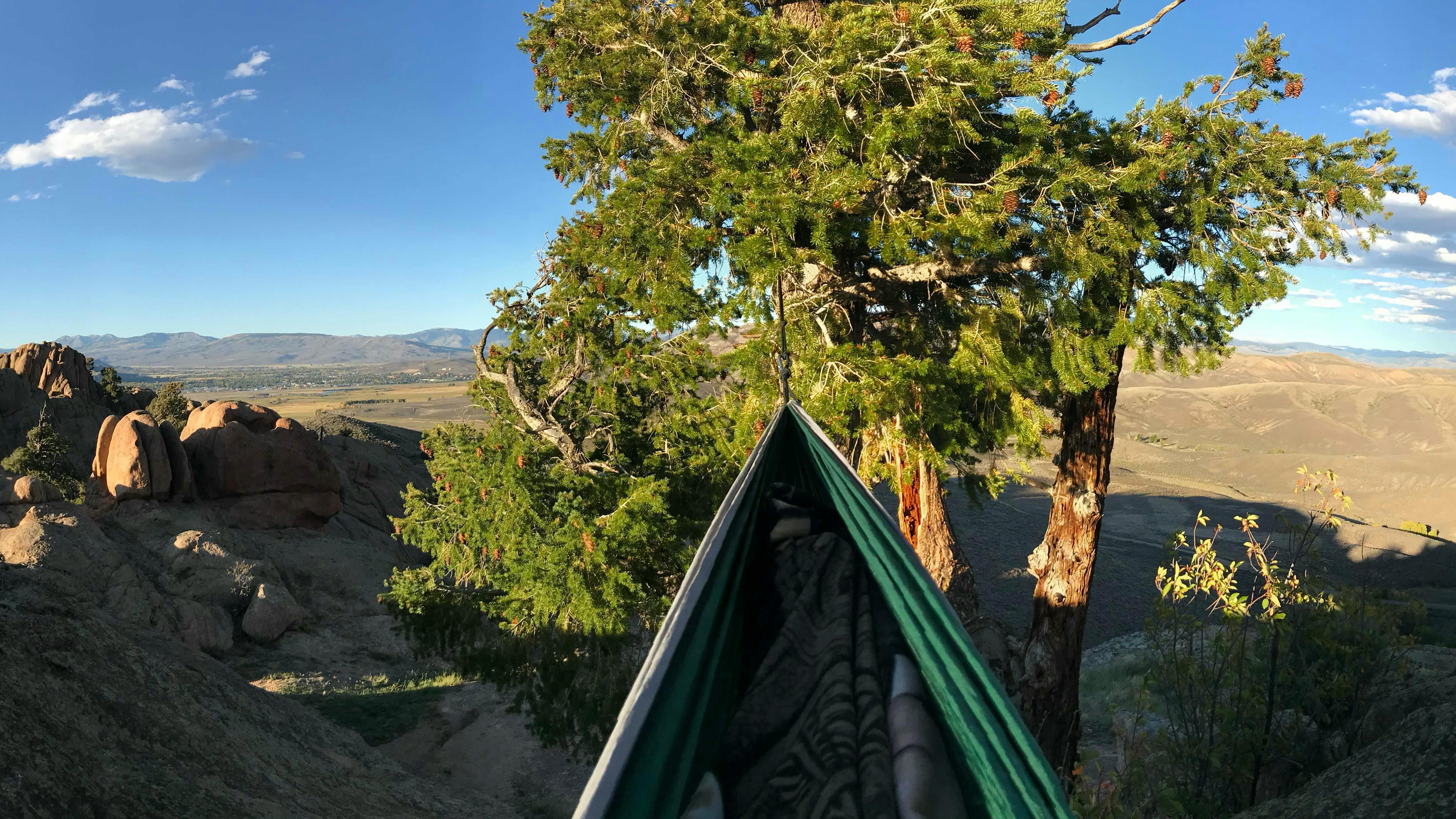 A green hammock is stretched between two trees and we look down the length of it towards an expansive view of interesting rock features.