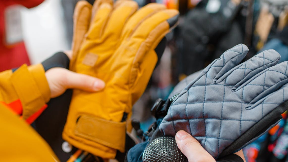 A person stands, holding one type of glove in each hand. There are more gloves in the background on a display at a store. 