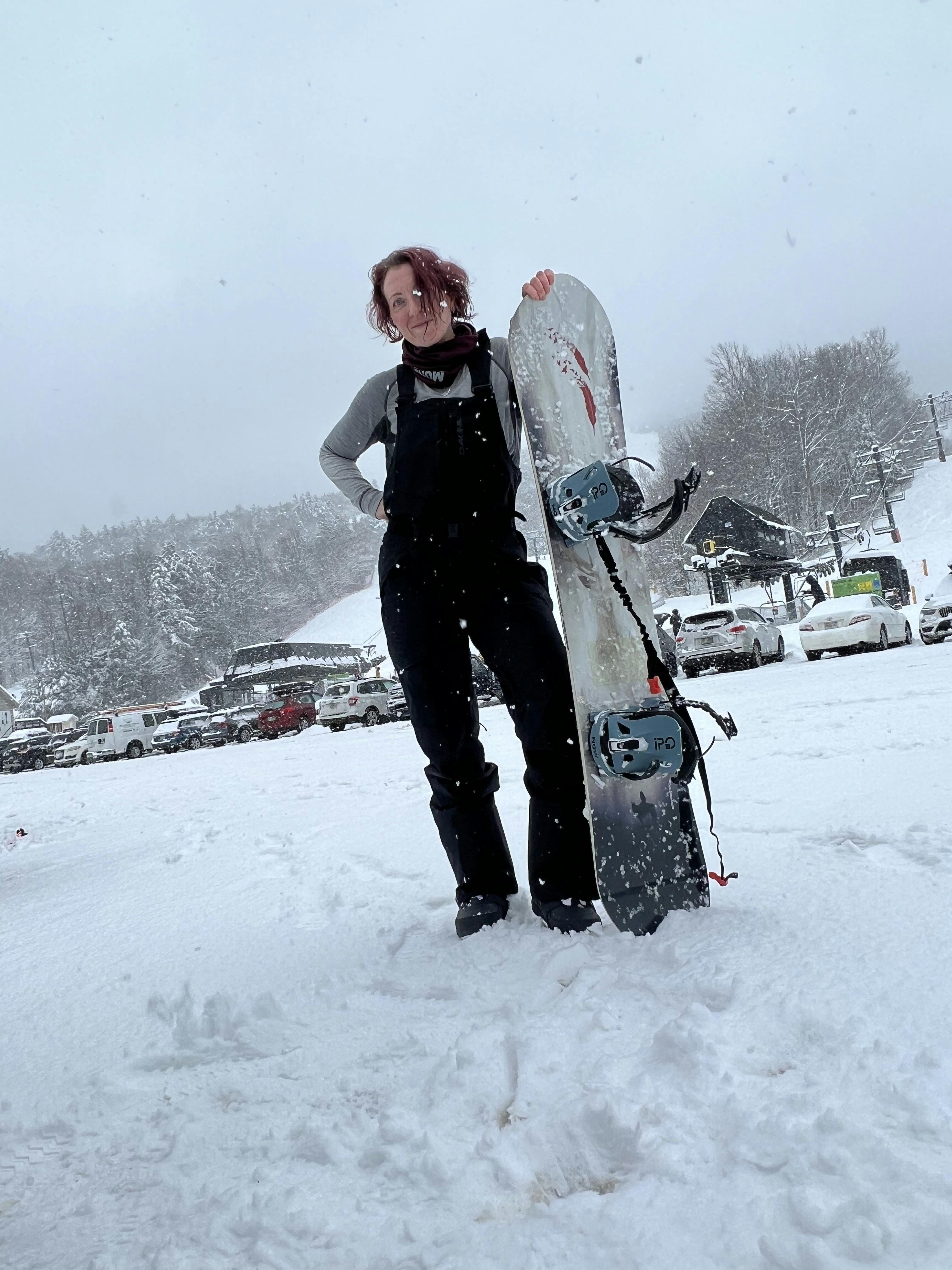 A snowboarder standing in the parking lot of a ski resort with her snowboard. 