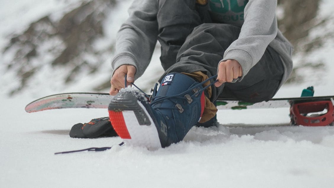 A snowboarder laces up his boots while sitting on his snowboard.