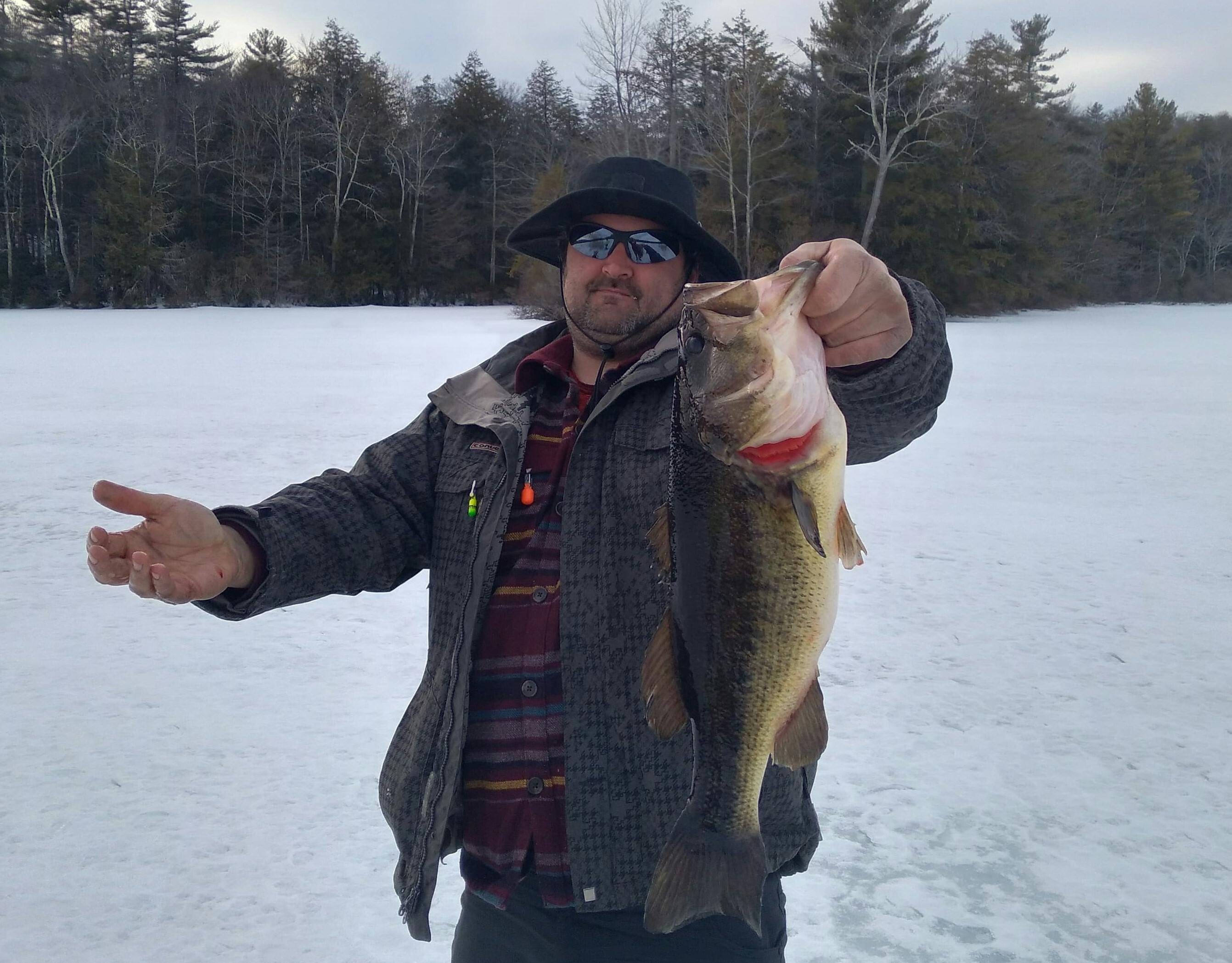 A man stands on ice and holds out a largemouth bass to the camera. He holds the fish by its mouth.