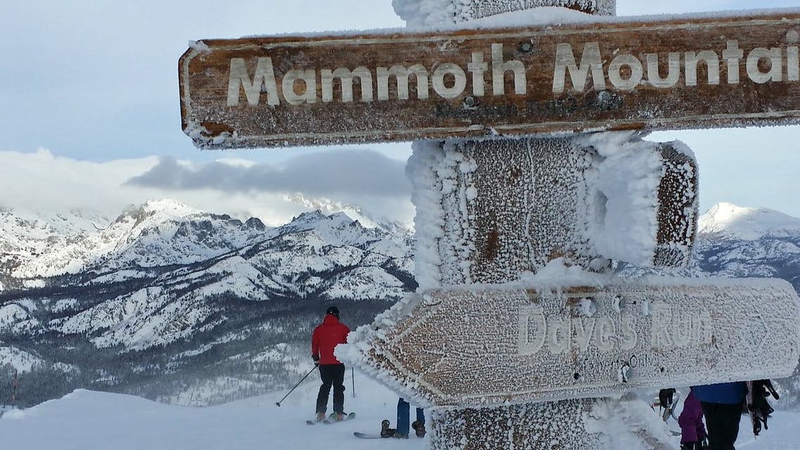 A snowy sign that says "Mammoth Mountain". There is a skier heading down the slopes in the background and several snowy mountains. 