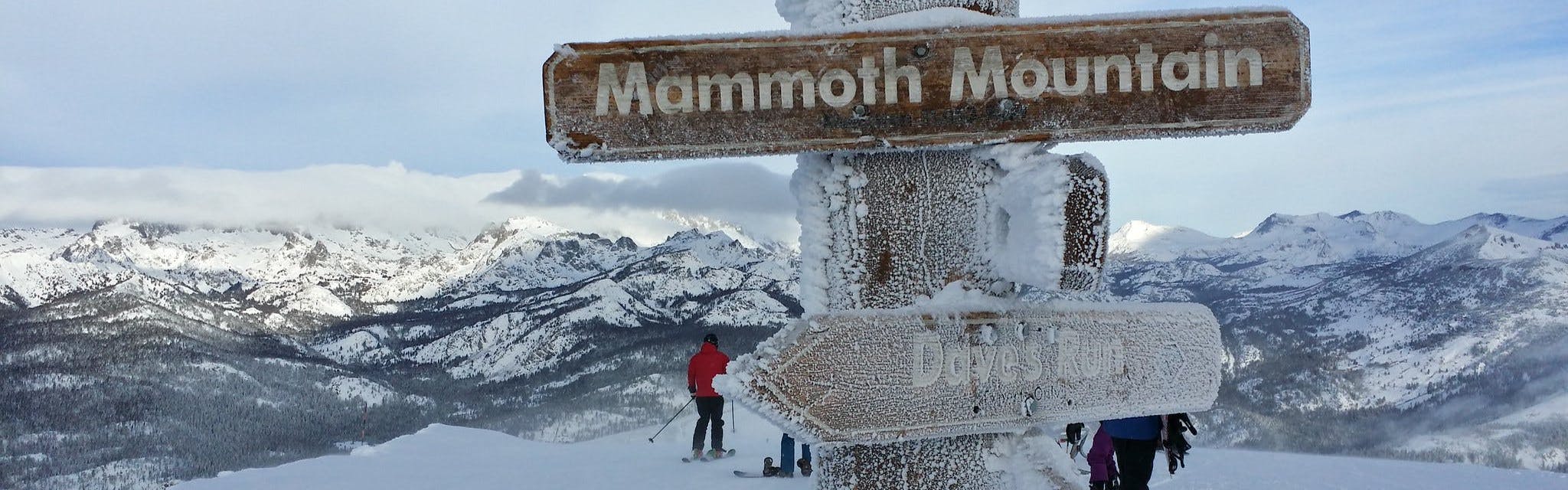 A snowy sign that says "Mammoth Mountain". There is a skier heading down the slopes in the background and several snowy mountains. 