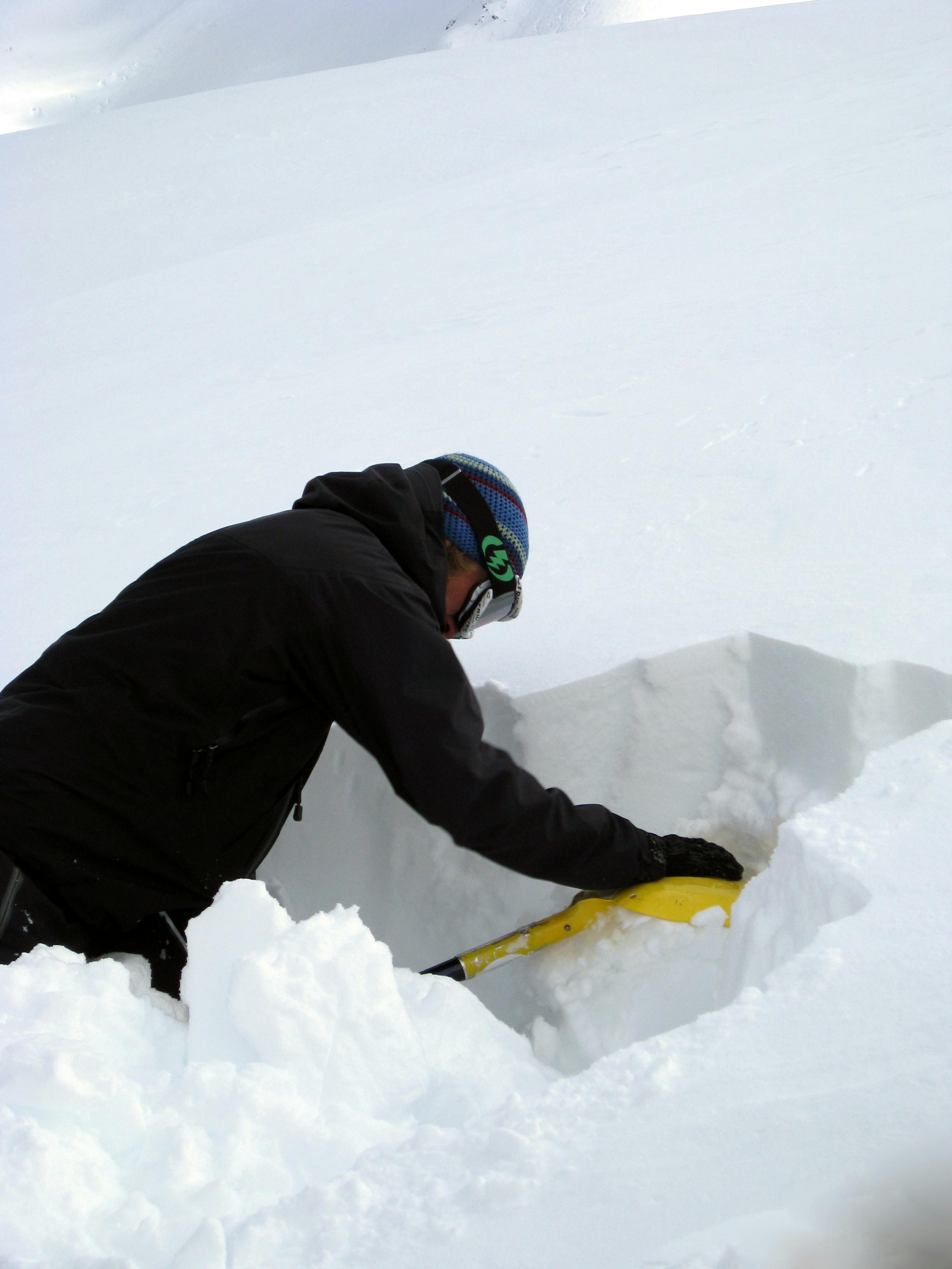 A man is using the head of a shovel to press down on some snow in a snow-pit for stability testing.