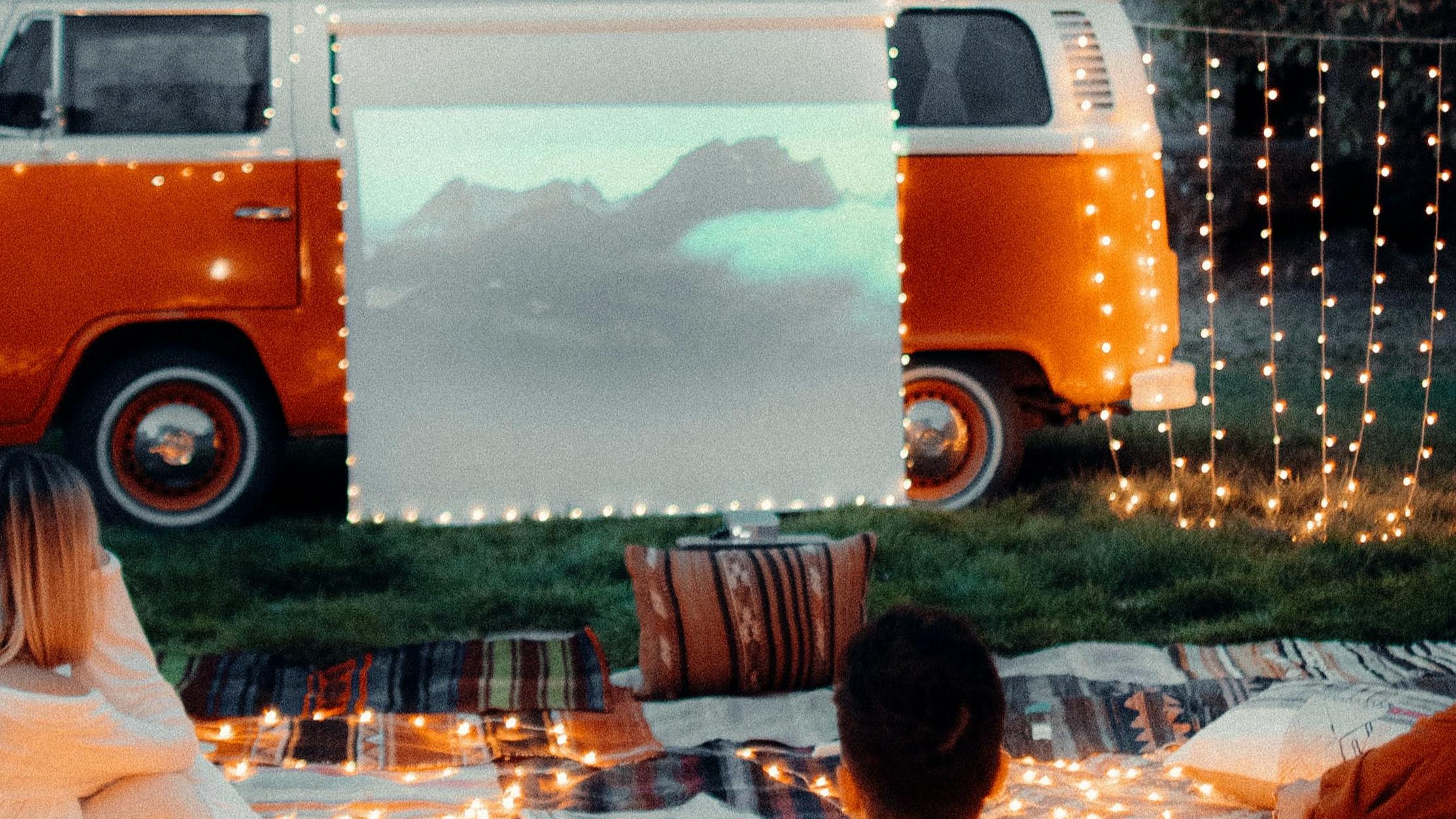 Several people watching a movie on a projector screen. There is an organge van that's holding up the projector screen and the movie watchers are sitting on picnic blankets. 