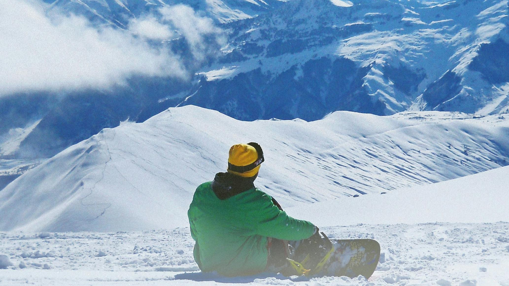 Snowboarder in a green jacket sitting on a snowy slope and looking out at the blue-tinged mountain range in the distance.