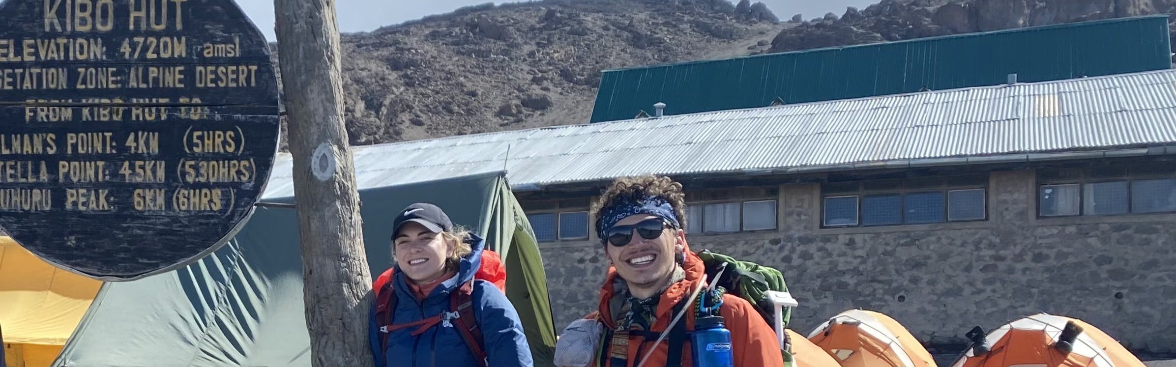 Two people stand at the bottom of a mountain with a sign indicating distances to different peaks. They are both wearing backpacking backpacks. 