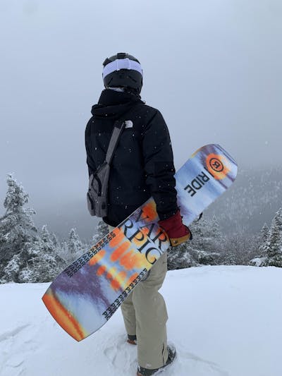 A man holding the Ride Warpig Snowboard at the top of a snowy ski run.
