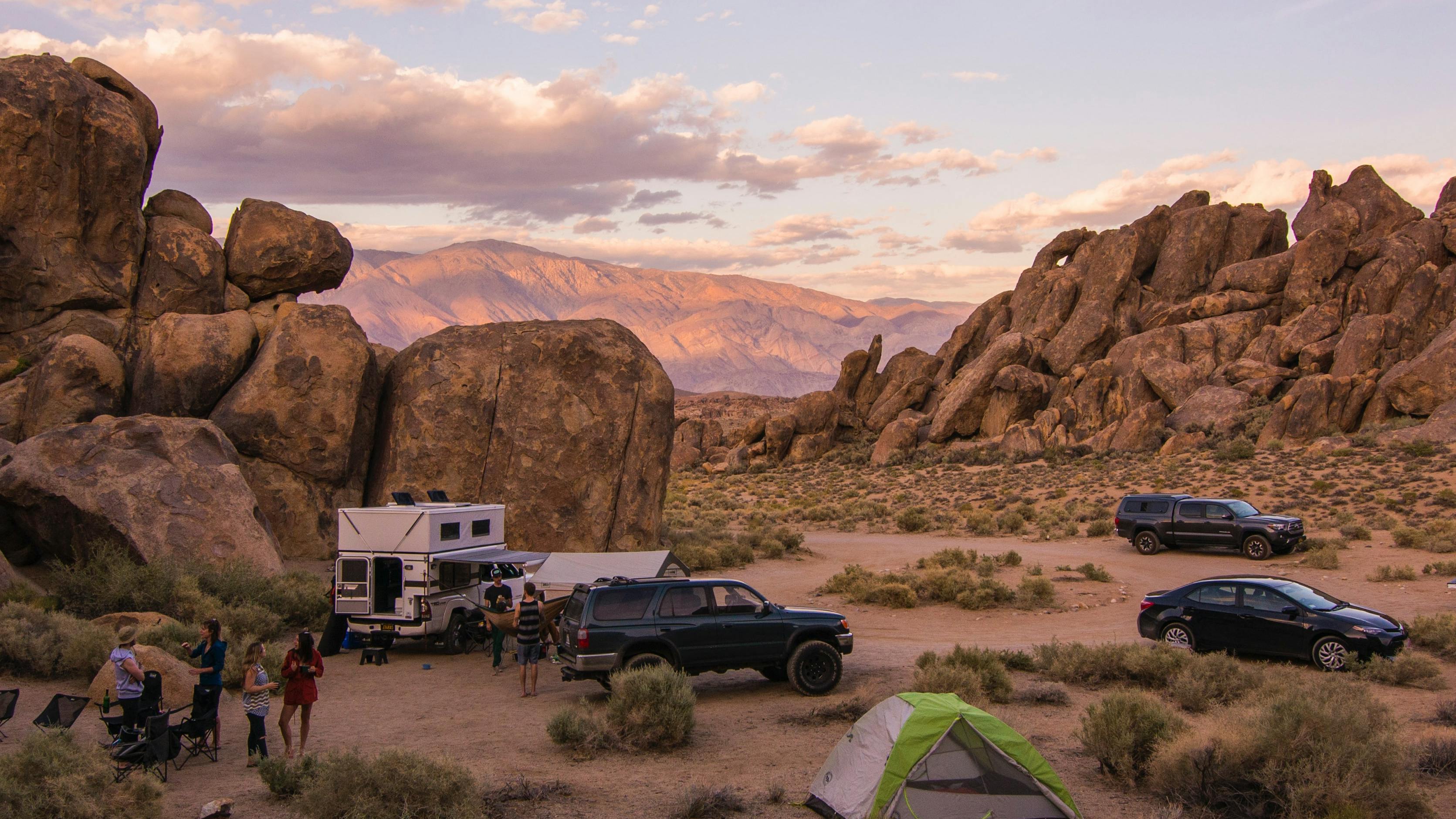 A campground with trucks, a camper, and tents in massive desert rock formations. The sky is pink and purple in the setting sun.