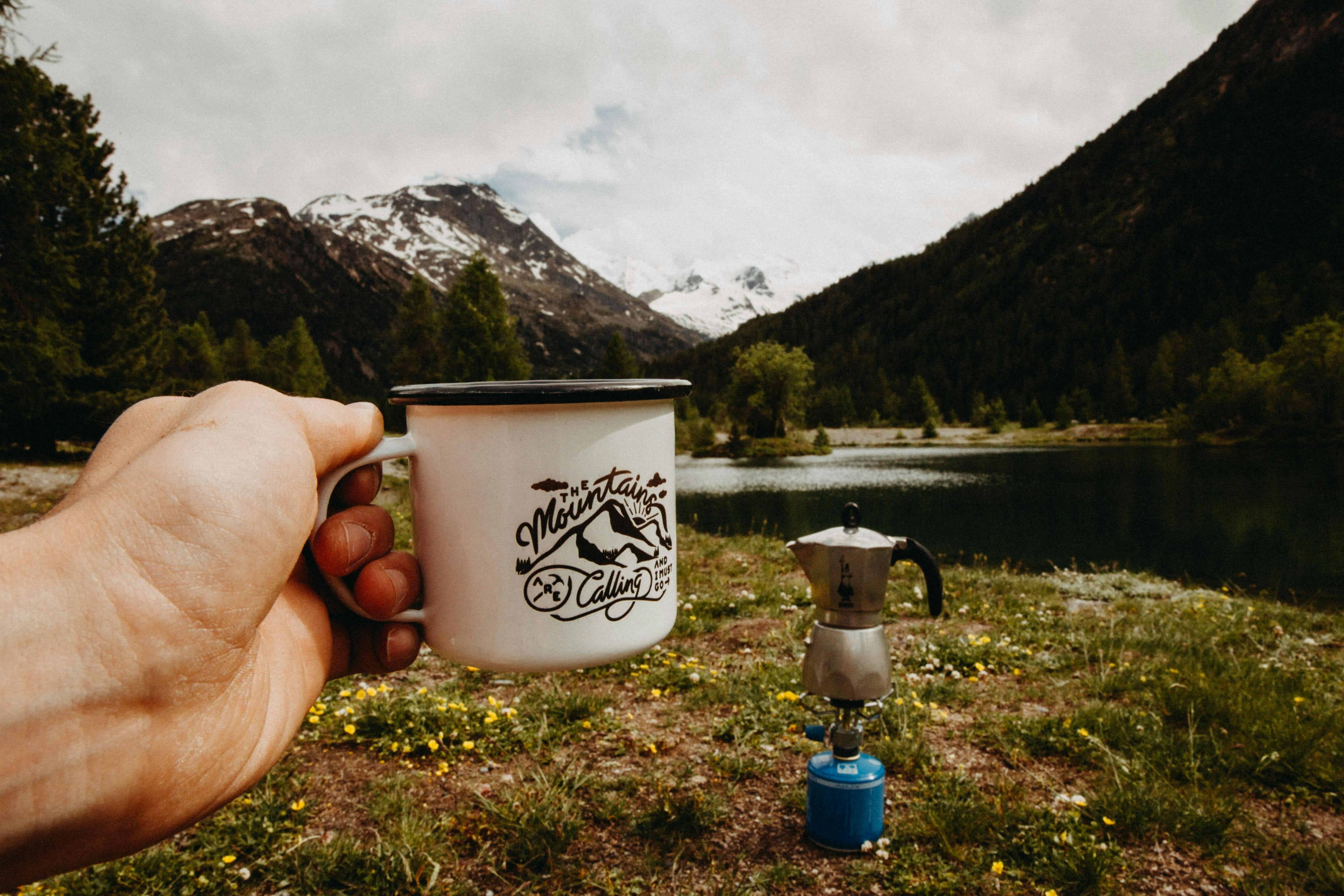Someone holds out a metal mug that reads "The Mountains Are Calling." In the meadow in front of them, they are brewing coffee in a Moka Pot next to a body of water with mountains rising in the distance. 