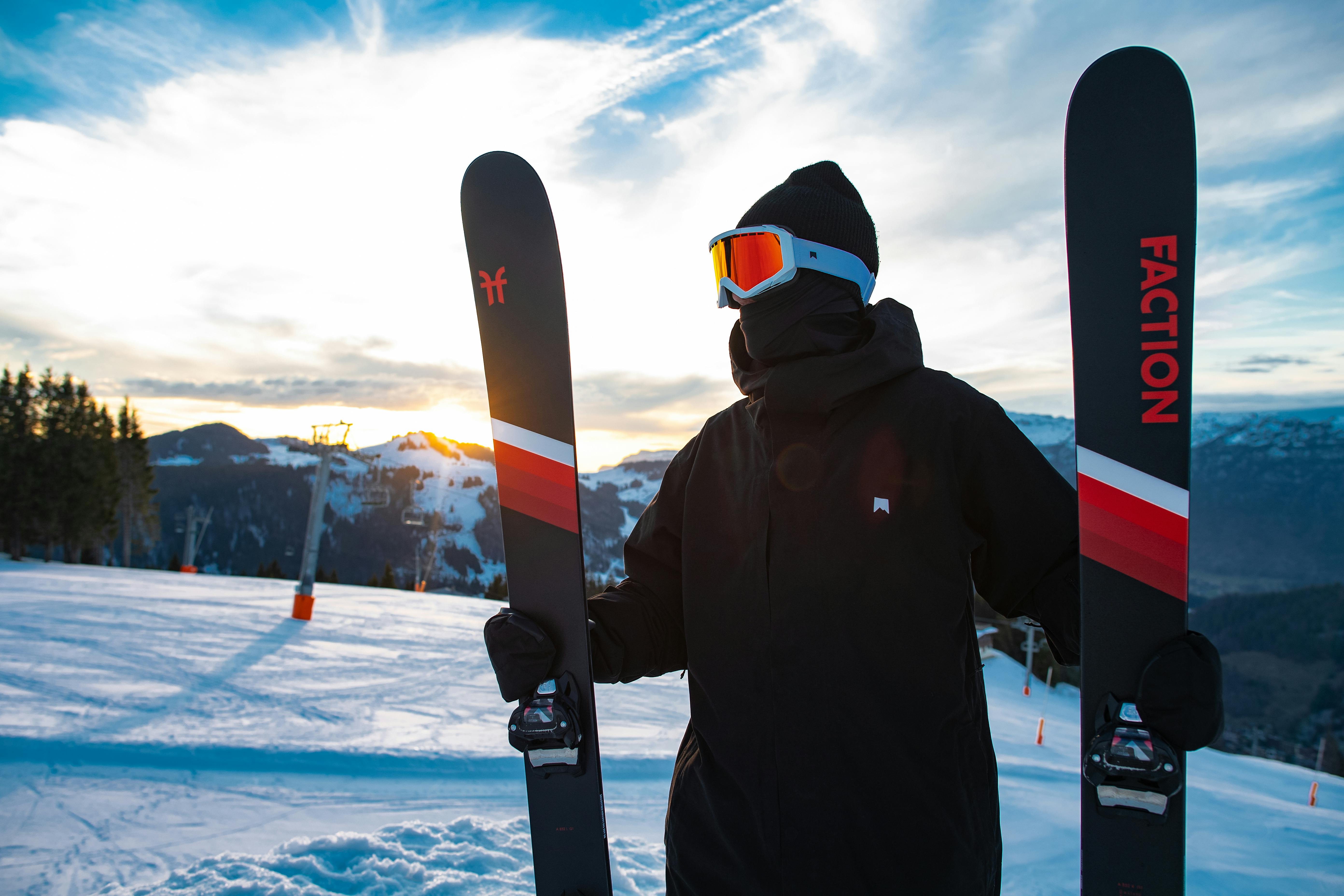 A man holding Faction skis on a slope with the sun setting in the background