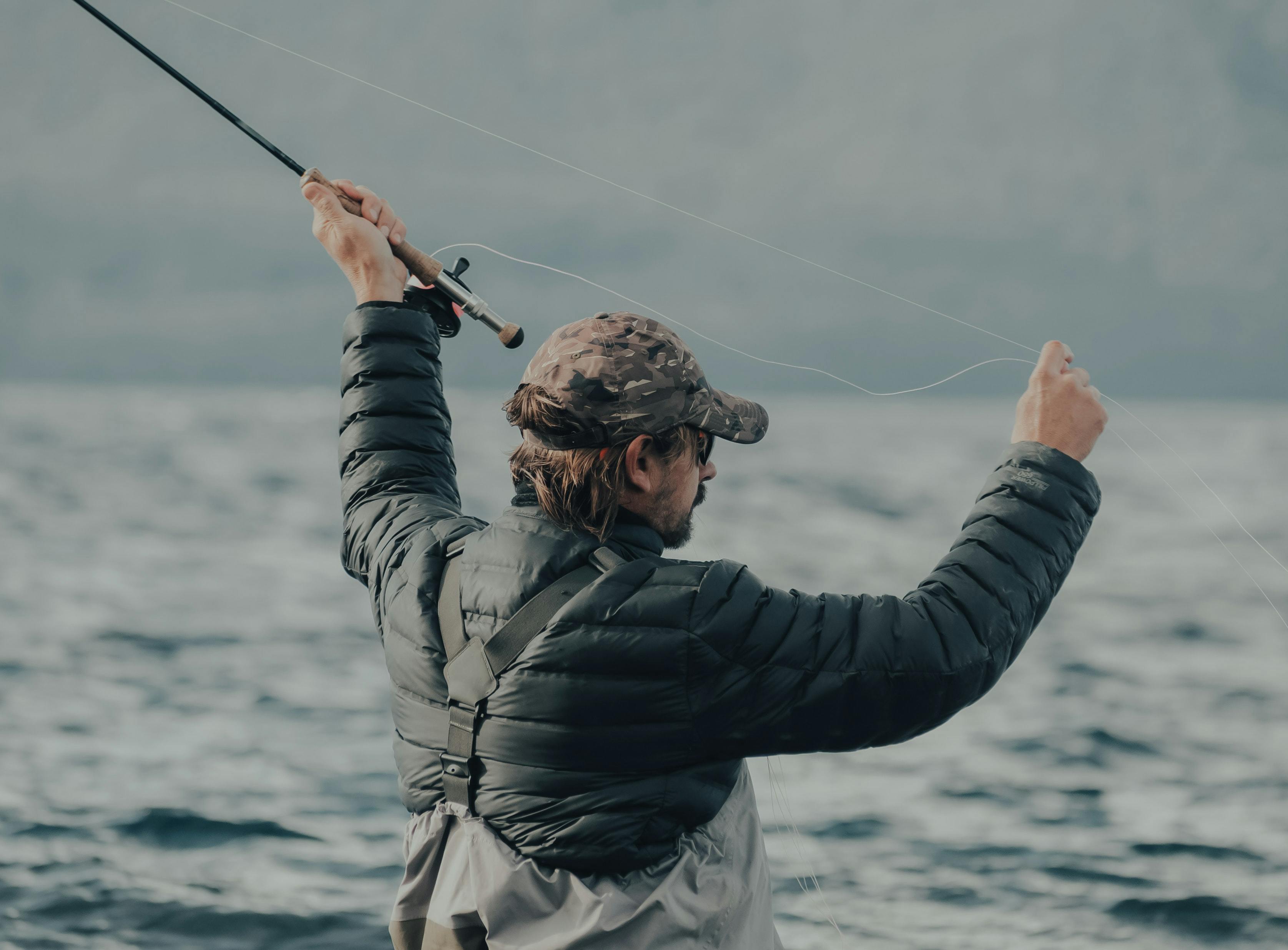 A man in waders, a puffy jacket, a baseball cap, and sunglasses casts his flyfishing line. 