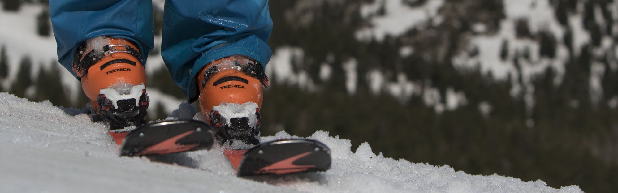 Close up photo of orange boots and orange and black skis on a ski slope. A mountain is visible in the background.