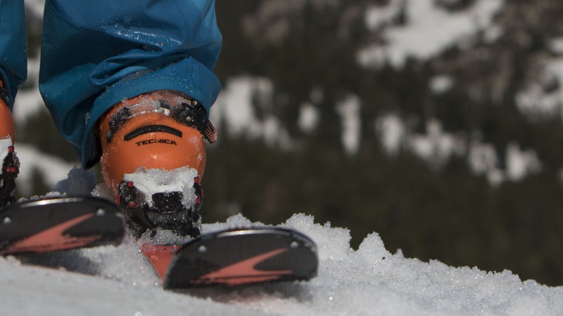 Close up photo of orange boots and orange and black skis on a ski slope. A mountain is visible in the background.