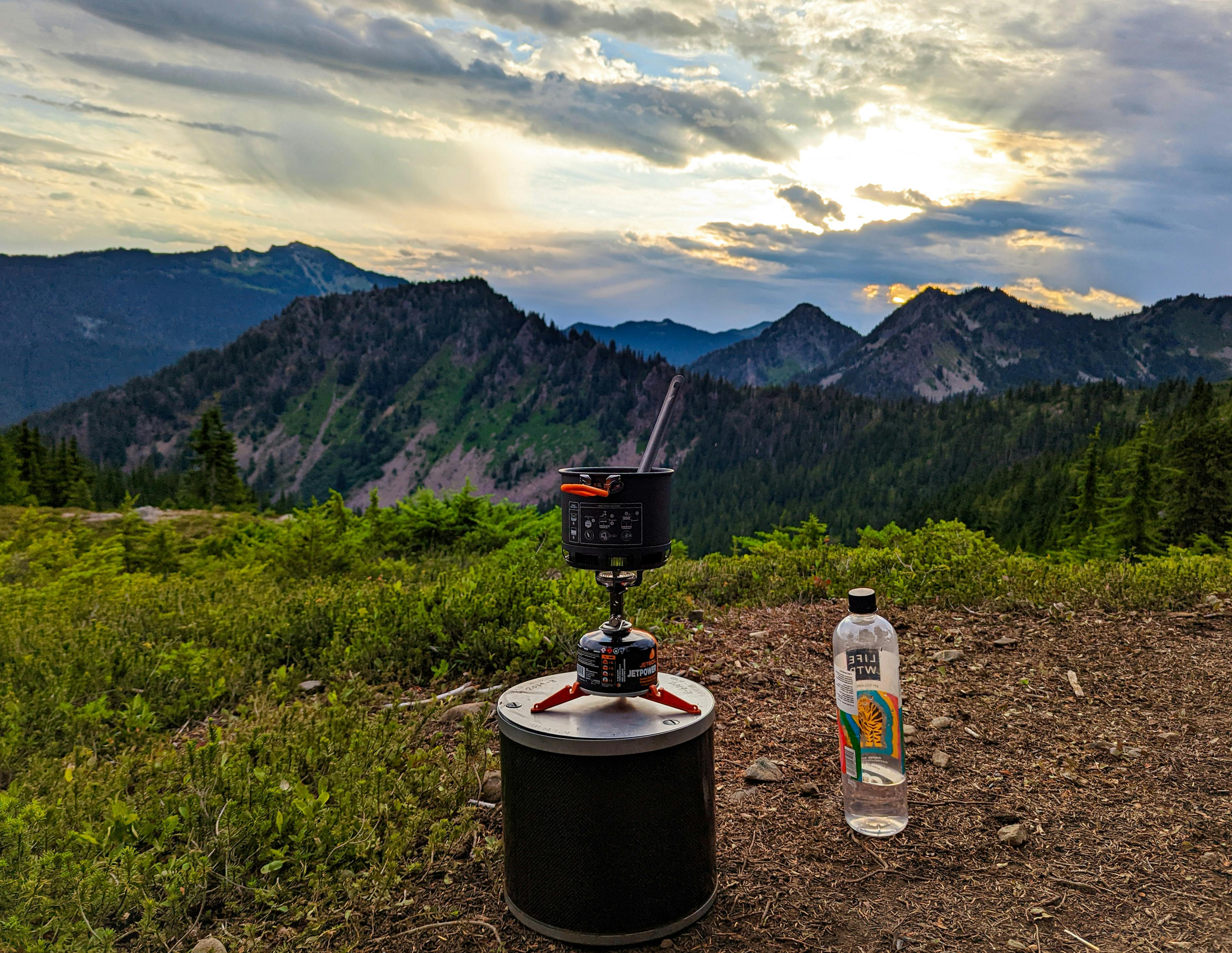 A camp stove on top of a bear canister. There are mountains in the distance. 