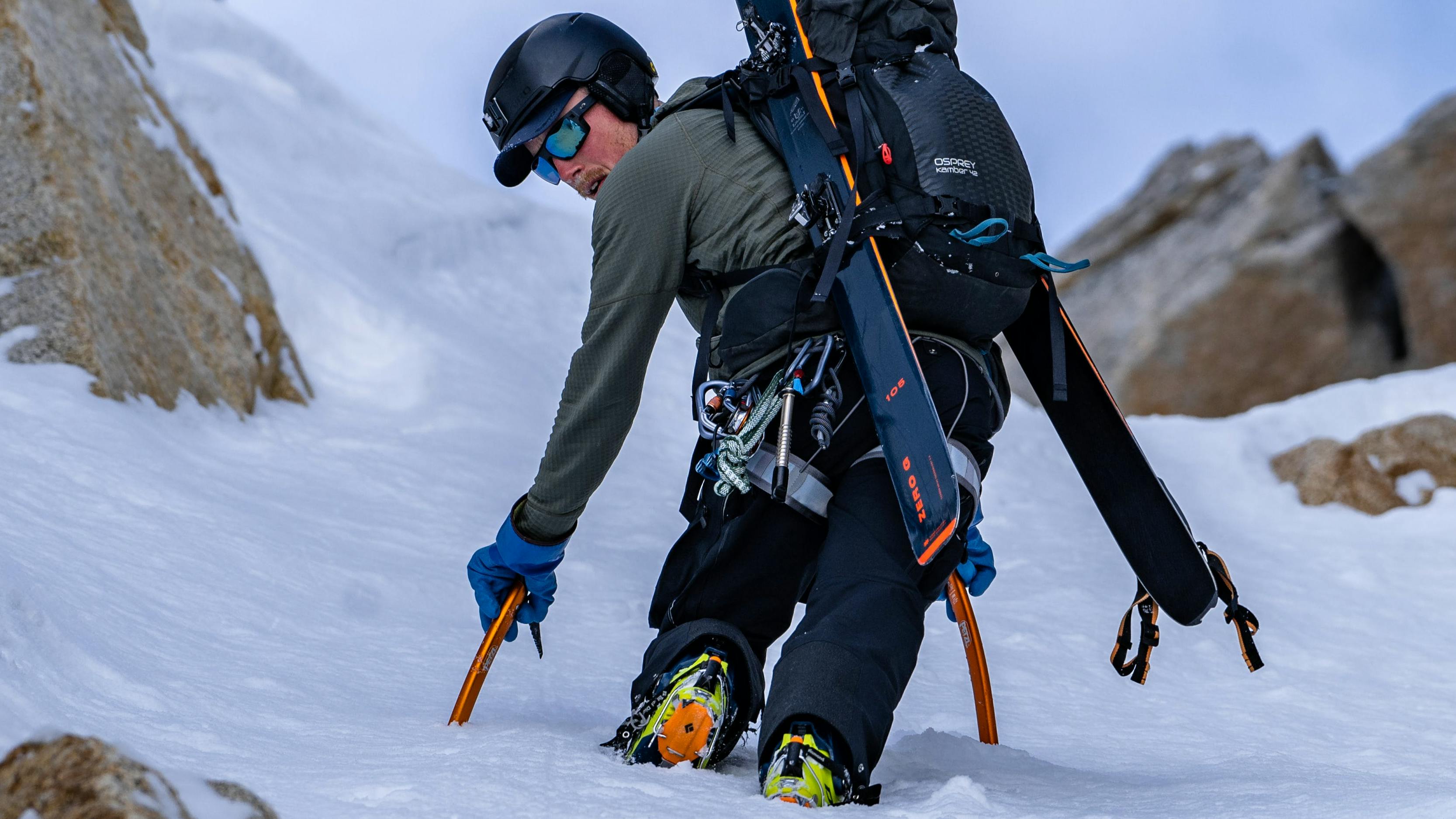A man ascends a steep chute with ice tools. His ski gear is on his backpack. 