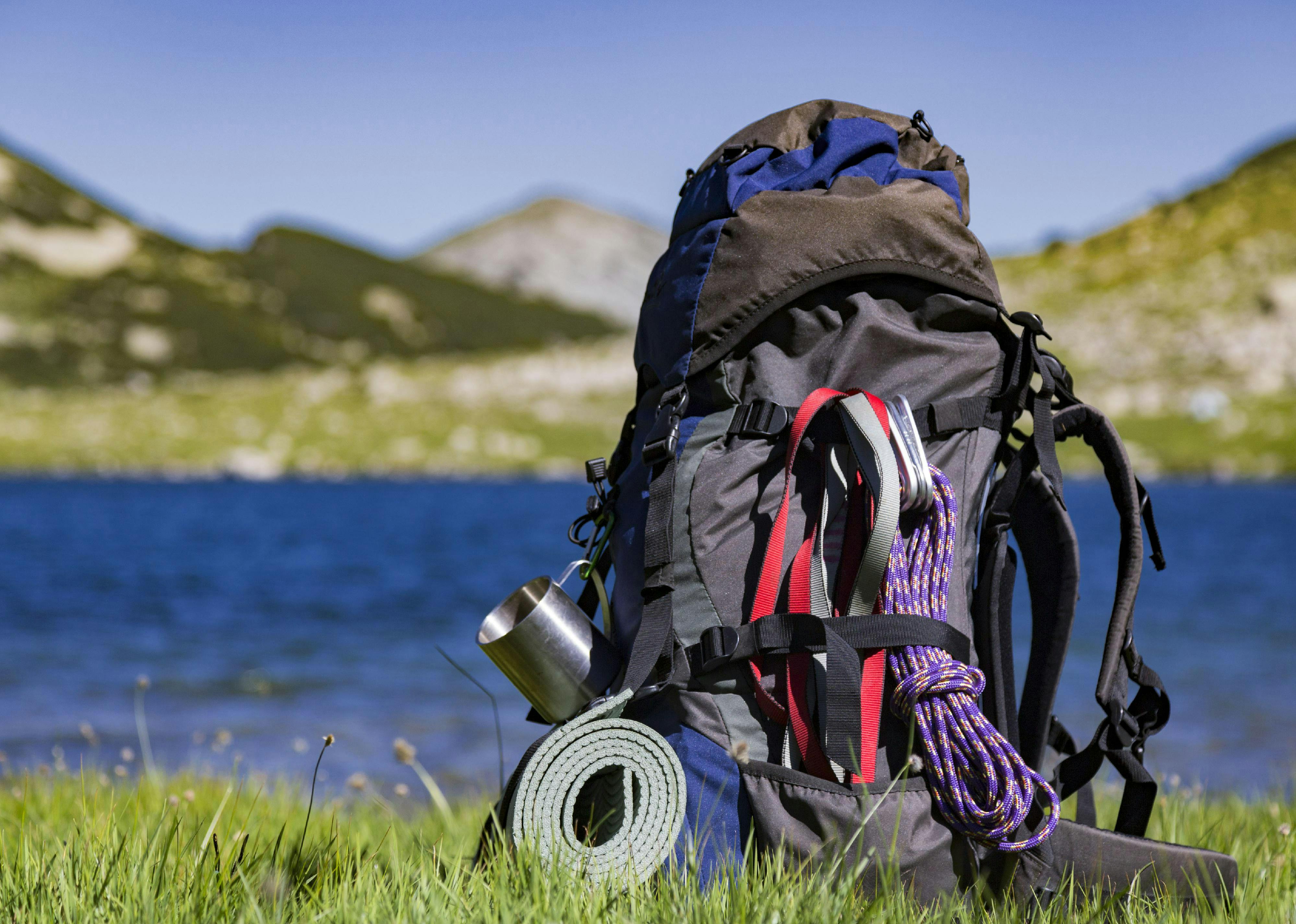 A backpacking pack rests next to a lake. On the outside of the pack, a metal mug, a sleeping pad, and rope are visible. 