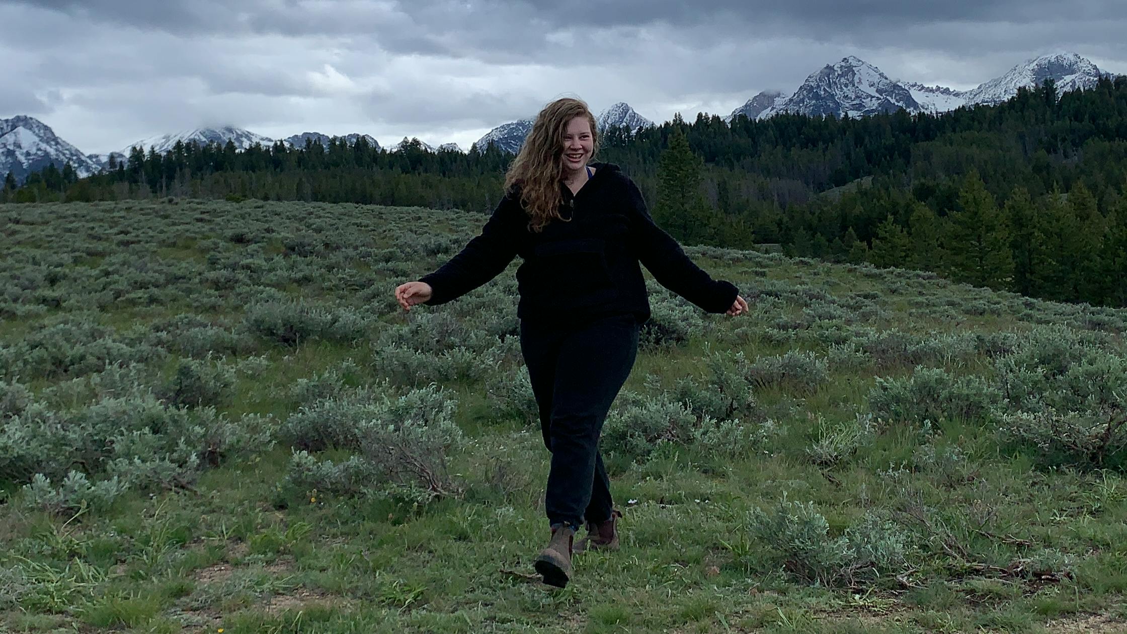 The author walks through a green field on a gray day. Snow-capped peaks line the background. 