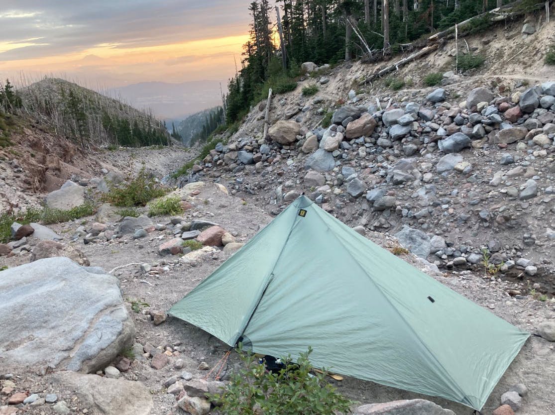 A tent is set up in a rocky area. A sunset is visible in the background.