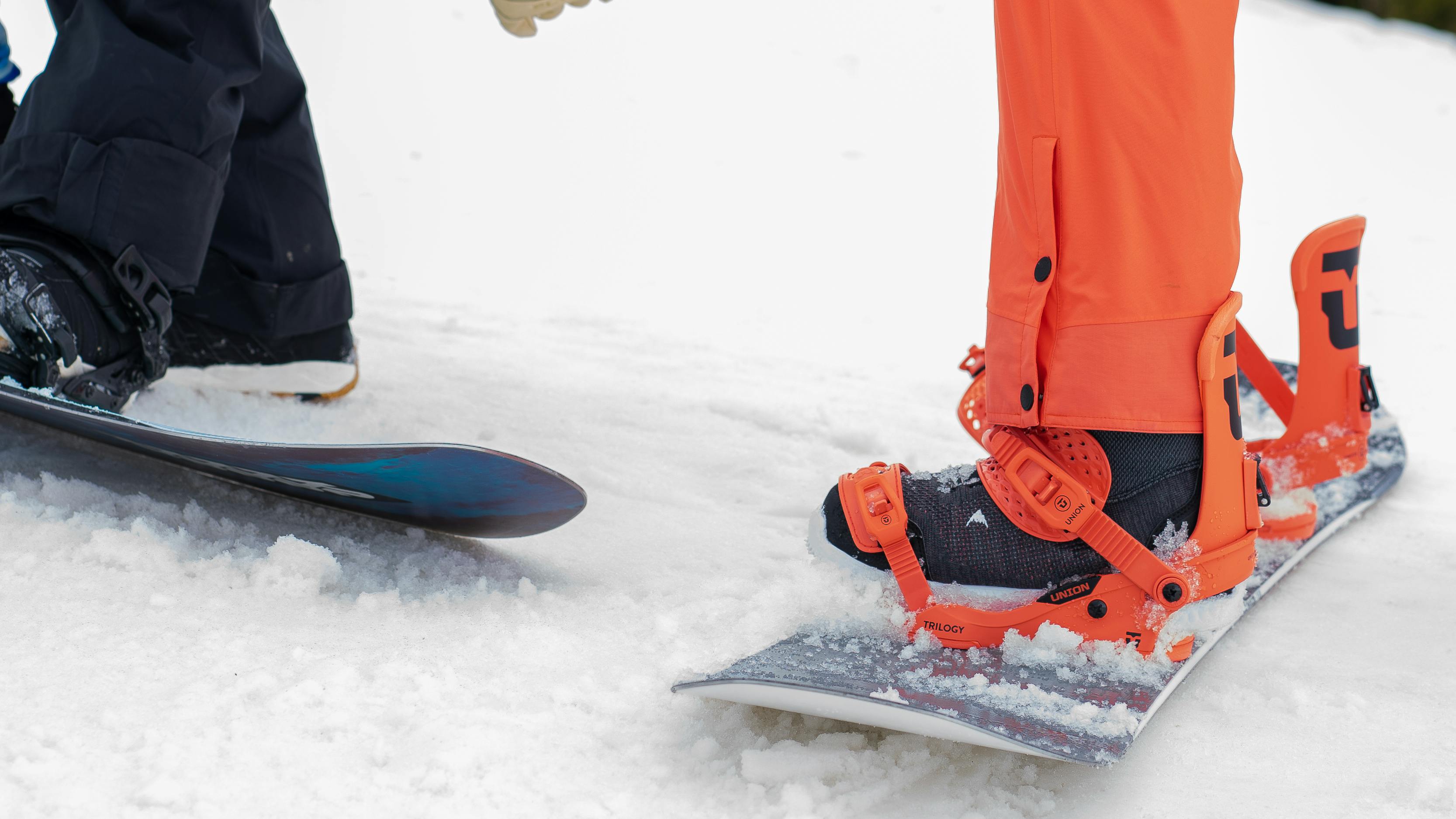 Two people on snowboards stand side by side, not yet strapped in.