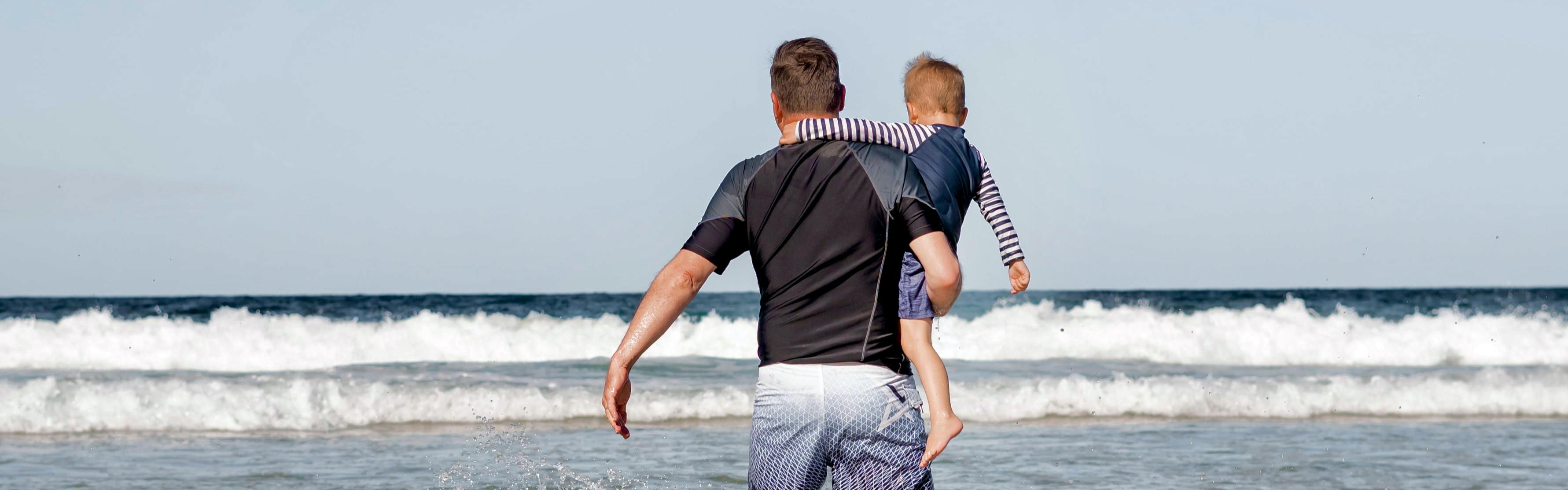 A man holds a young boy on his hip and walks toward the ocean. They both wear board shorts and sun shirts.