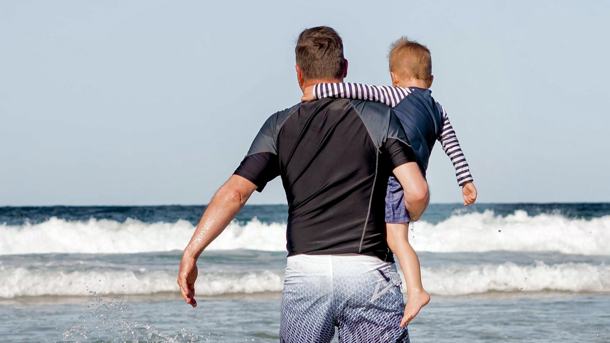 A man holds a young boy on his hip and walks toward the ocean. They both wear board shorts and sun shirts.