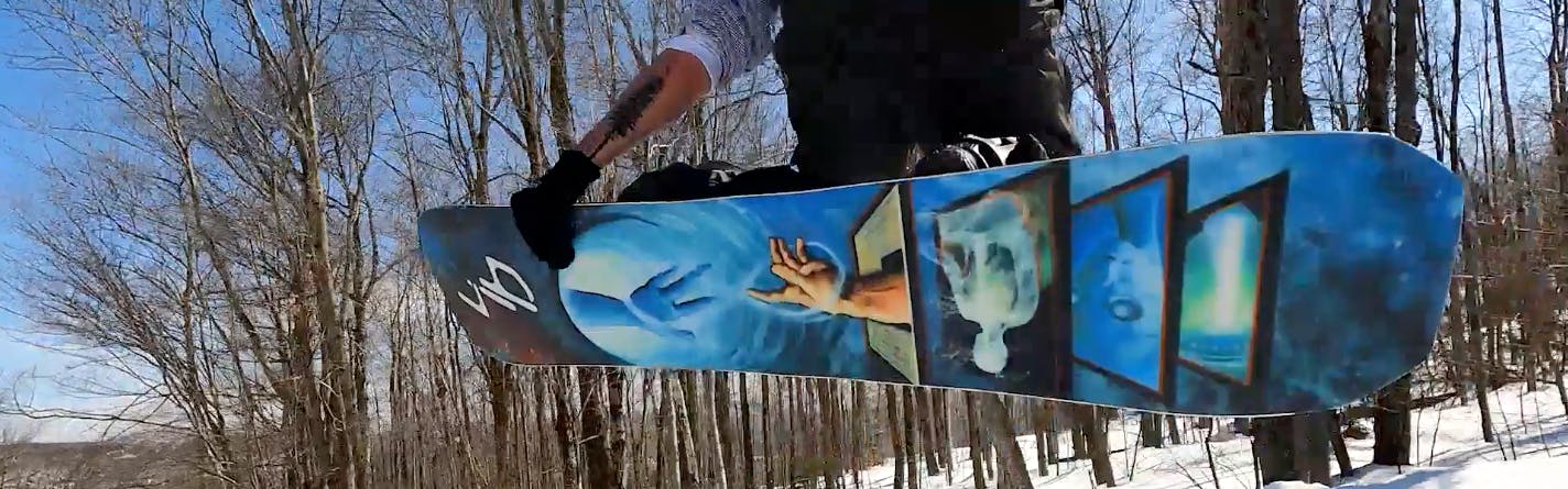 Curated expert Colby Henderson jumping with the Lib Tech Golden Orca snowboard, the bottom of the board fully visible as he grabs hold of it with his left hand in the air