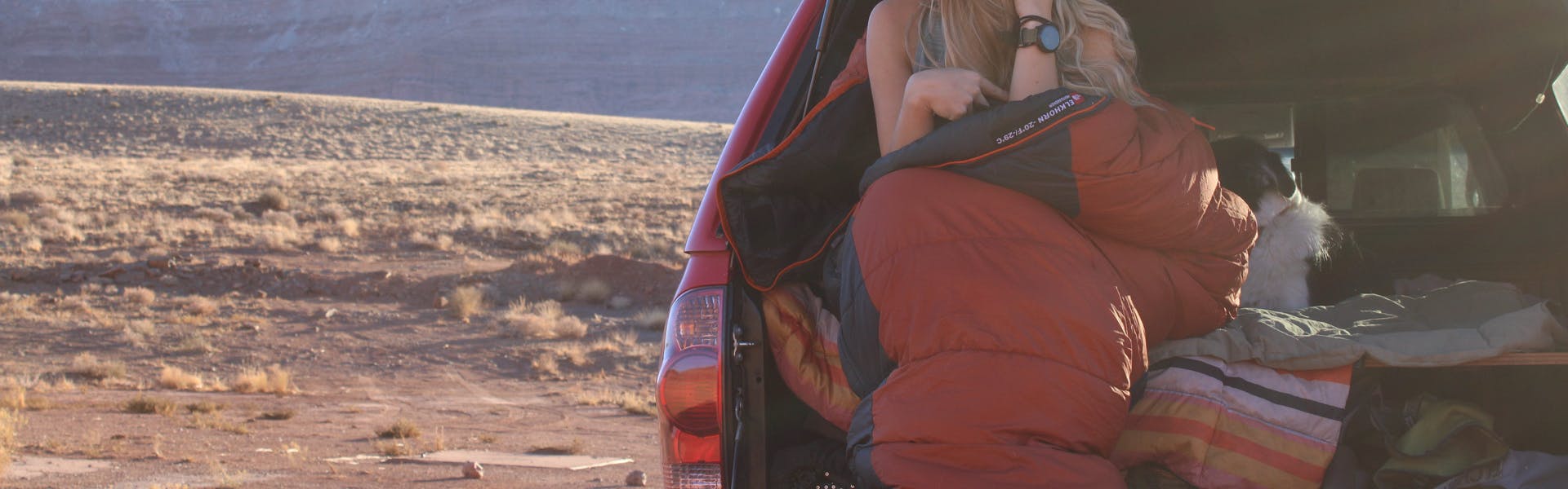 A woman sitting on a tailgate with a sleeping bag around her. 