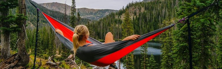 A woman reclines in a red and grey hammock overlooking trees and water