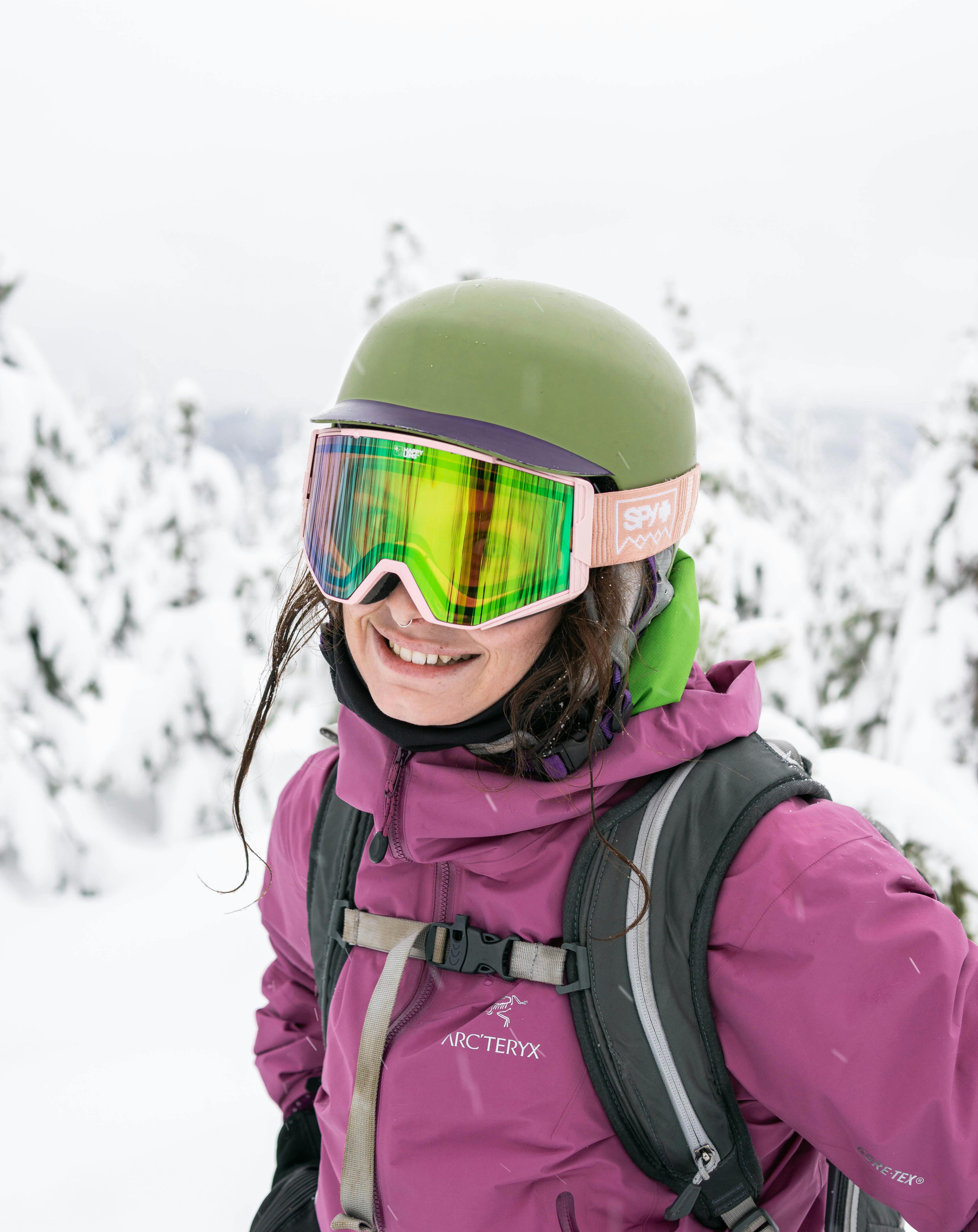 A woman in a pink jacket smiles at the camera while wearing goggles and a helmet. 