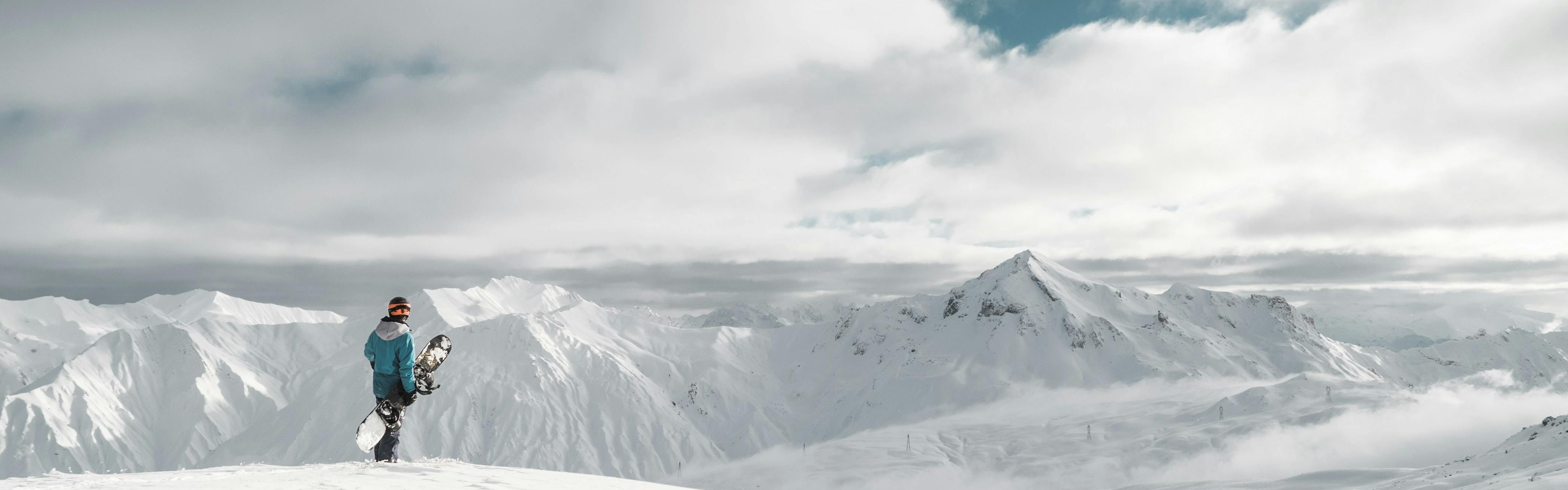 A snowboarder stands with his board on top of a snowy mountain range.