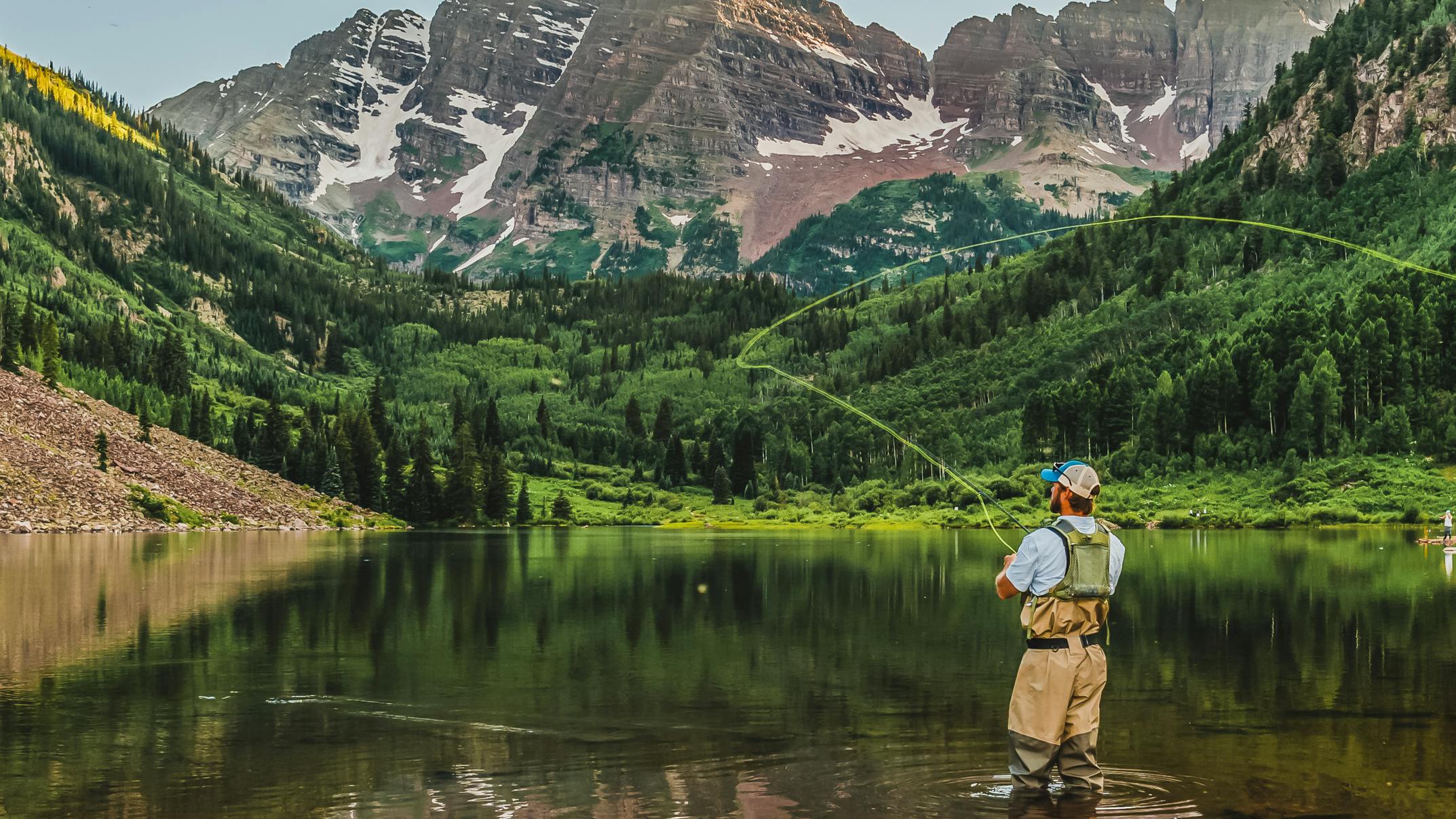 A man stands in an alpine lake and casts his fly rod. 