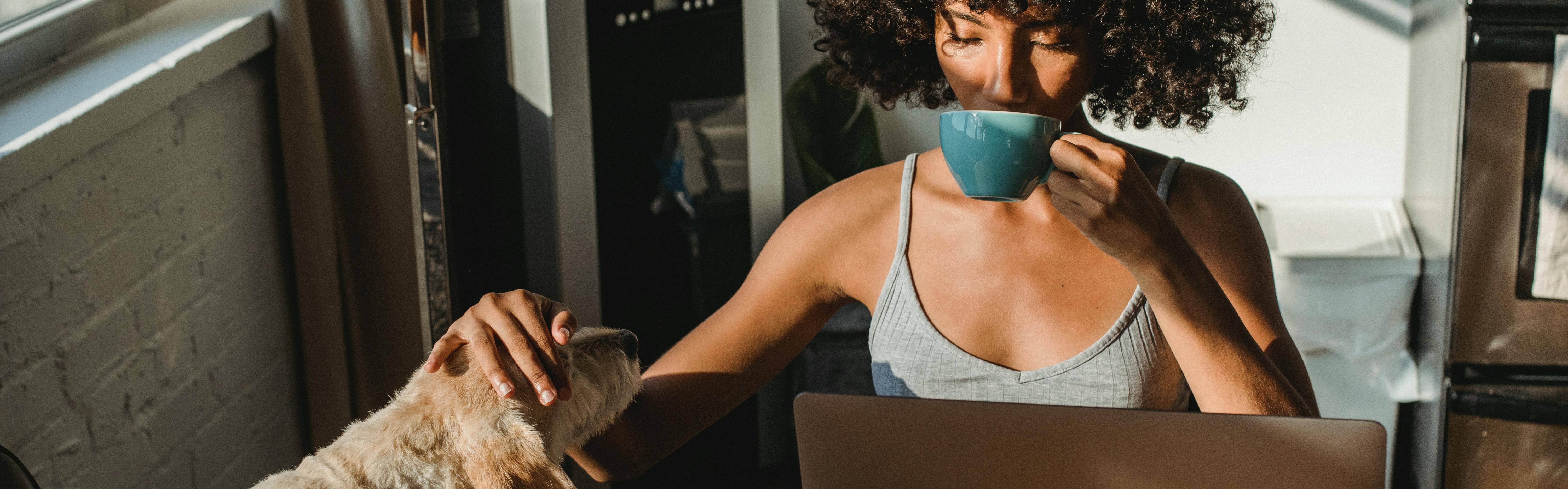 A woman drinking coffee in front of her laptop while she pets a dog. 