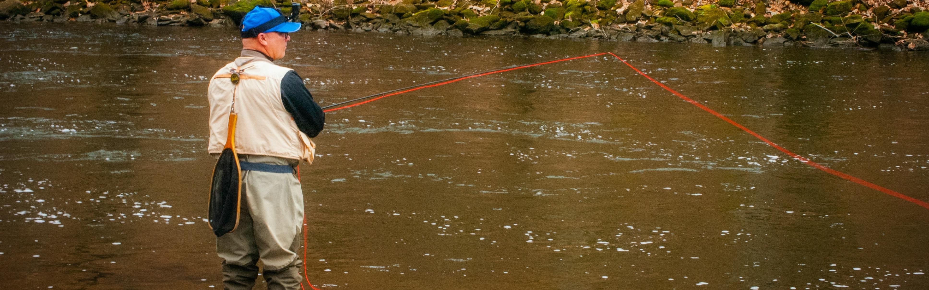 A man with waders and a fishing vest stands in a river. He is casting his fly rod and has a fishing net attached to his back.