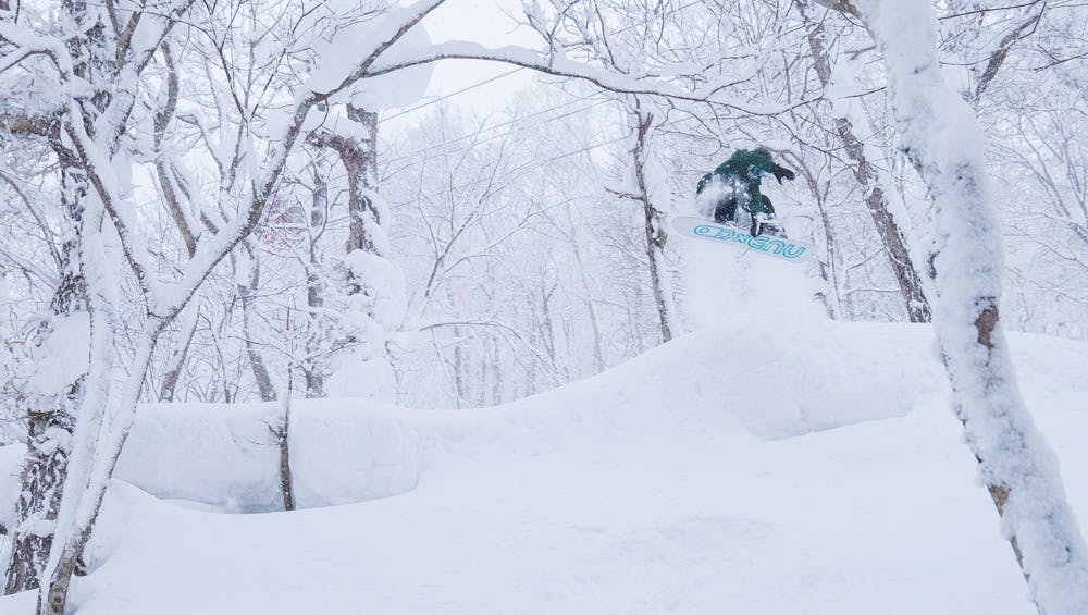 A snowboarder jumping in a very snowy wooded area. 