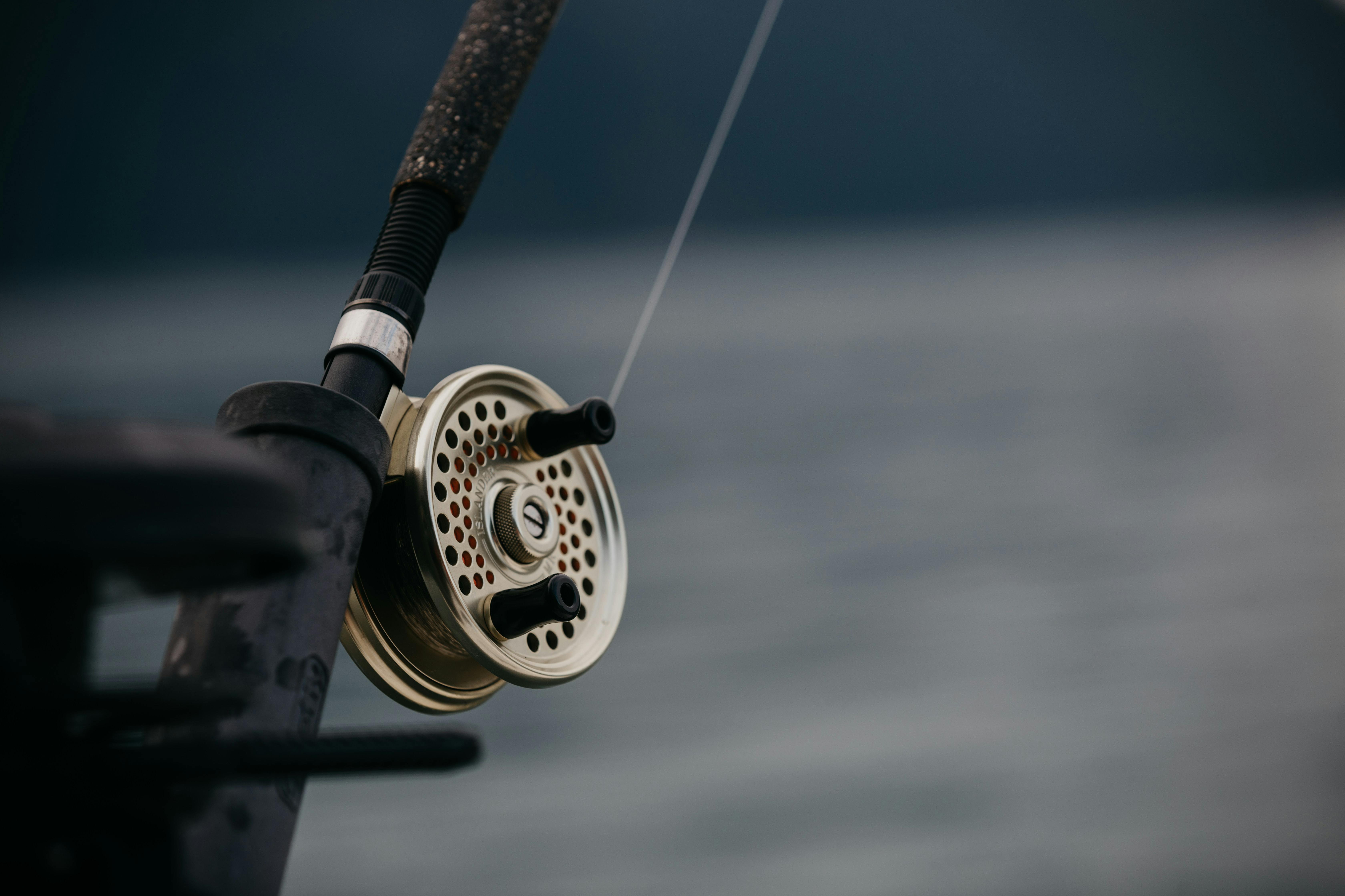 A fly fishing reel attached to a rod with a body of water in the background. 